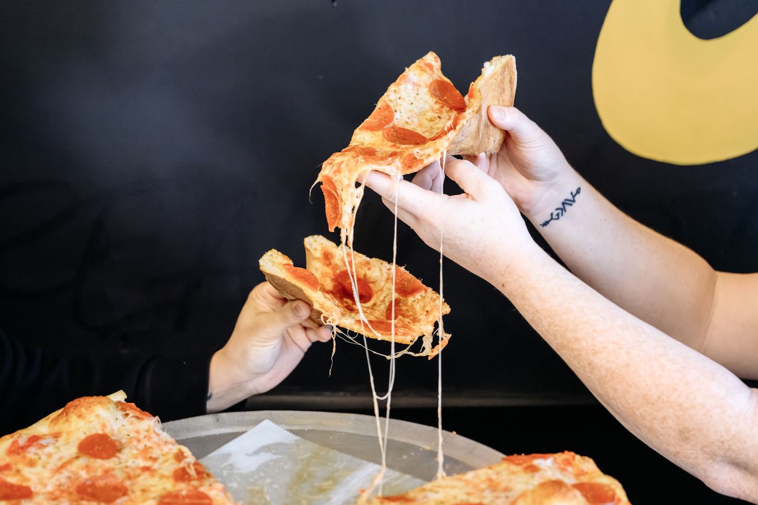 Hands pull apart cheesy pepperoni pizza slices with long stretchy mozzarella cheese over a restaurant table.