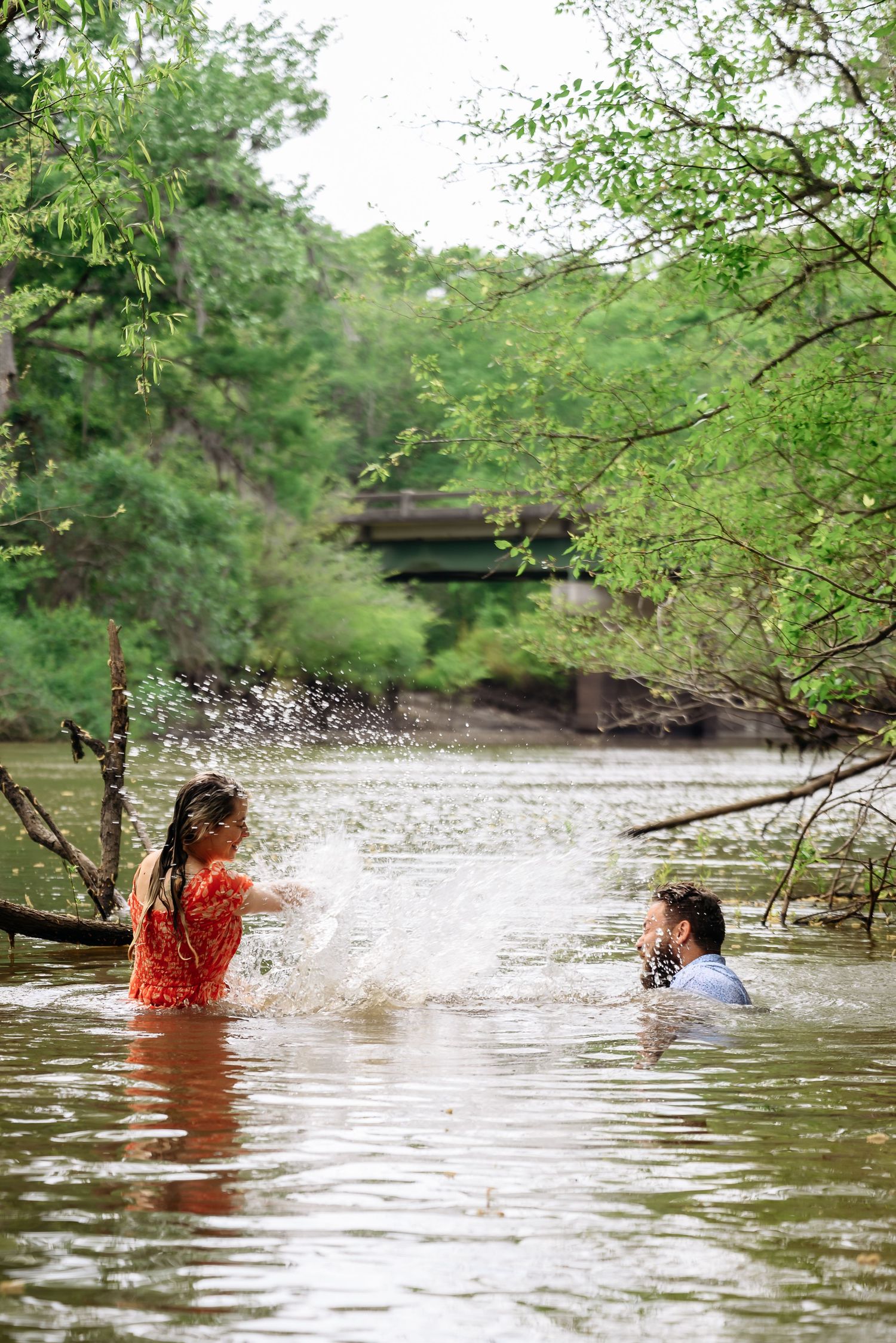 Couple playfully splashing water in a river surrounded by lush green trees during an outdoor photo session.
