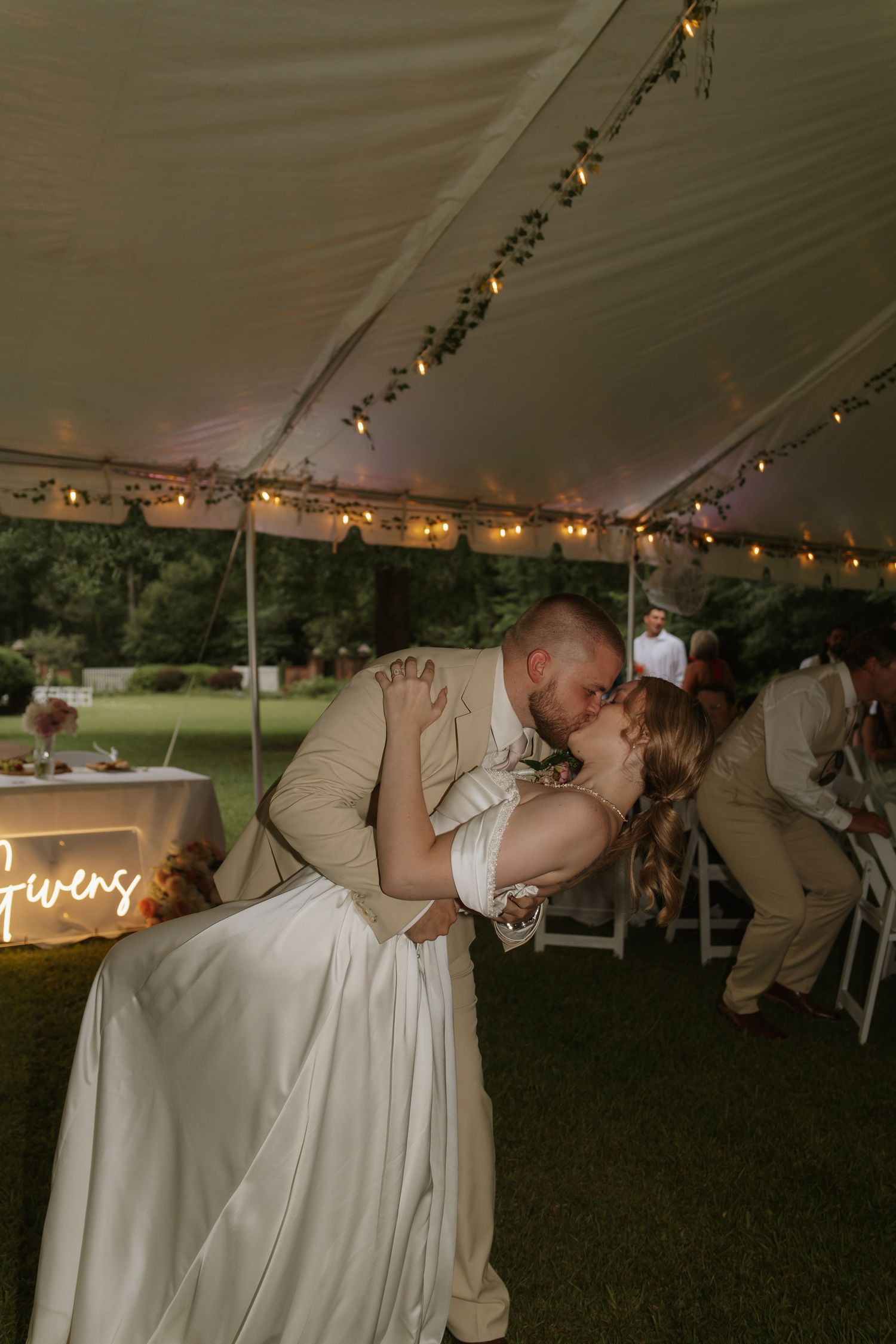 Bride and groom share a romantic dip kiss under a white tent with string lights at an outdoor wedding reception.
