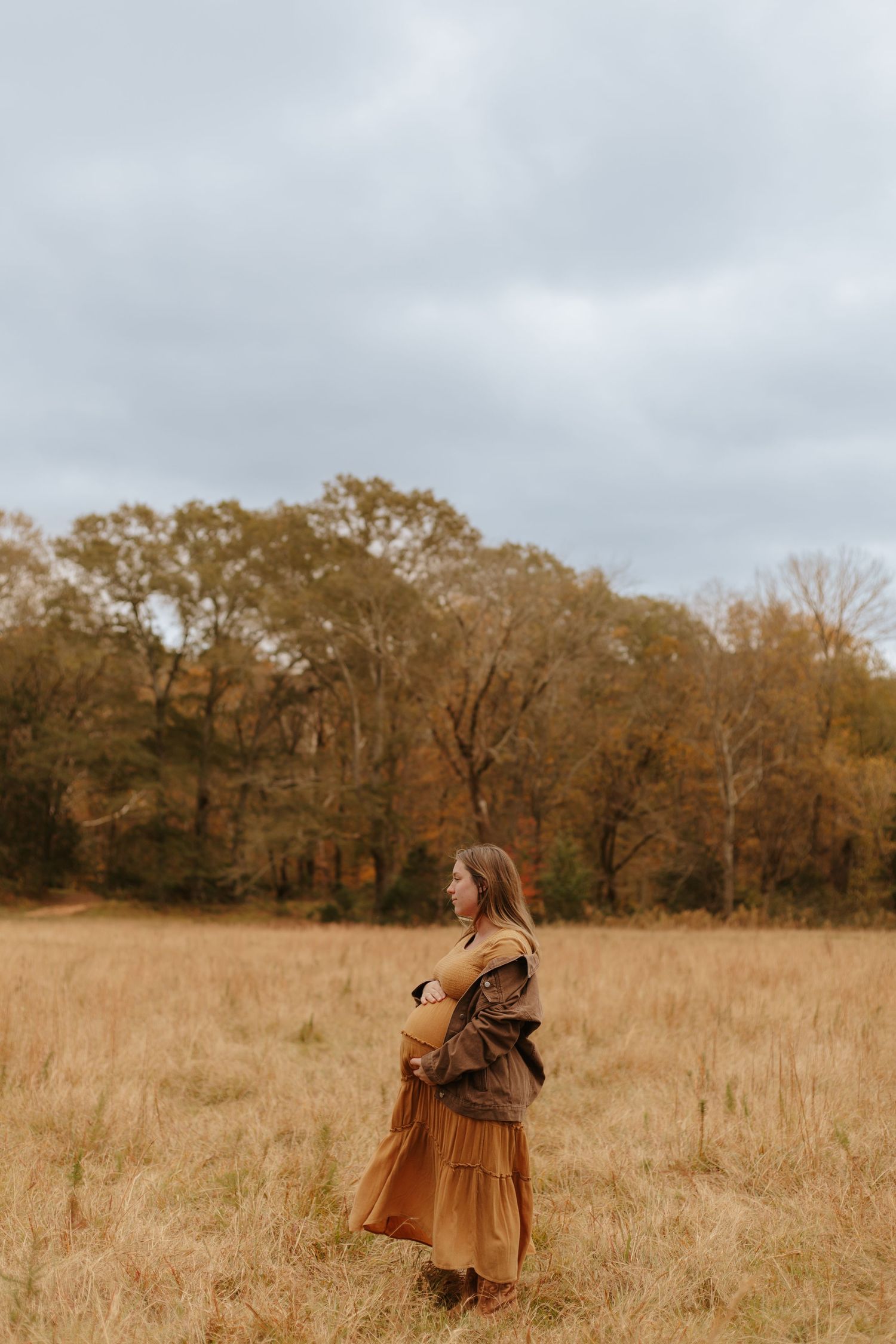 Woman in brown maxi dress and jacket standing in golden autumn field under overcast sky.