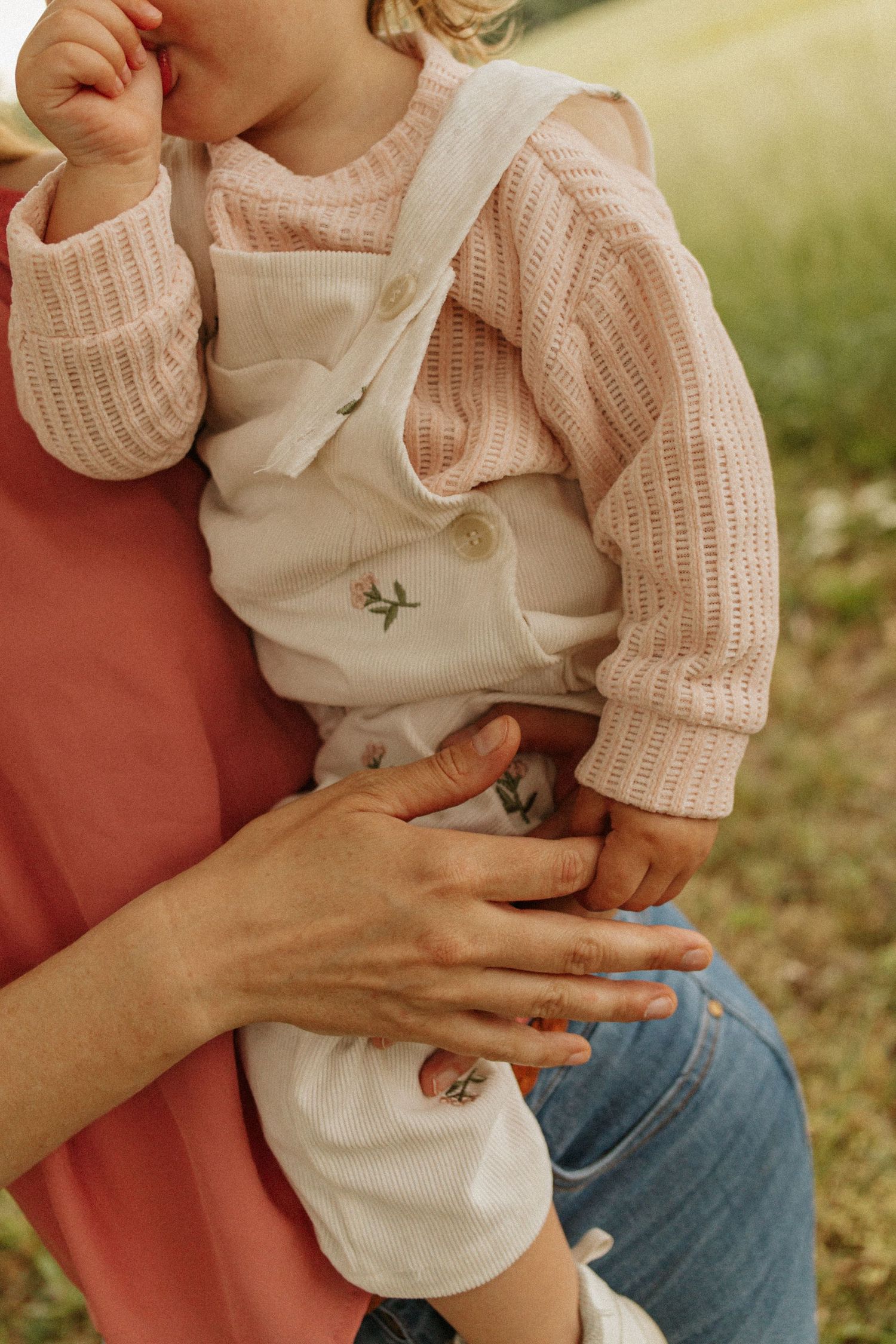 Mother holding baby in floral overalls and pink knit sweater outdoors.