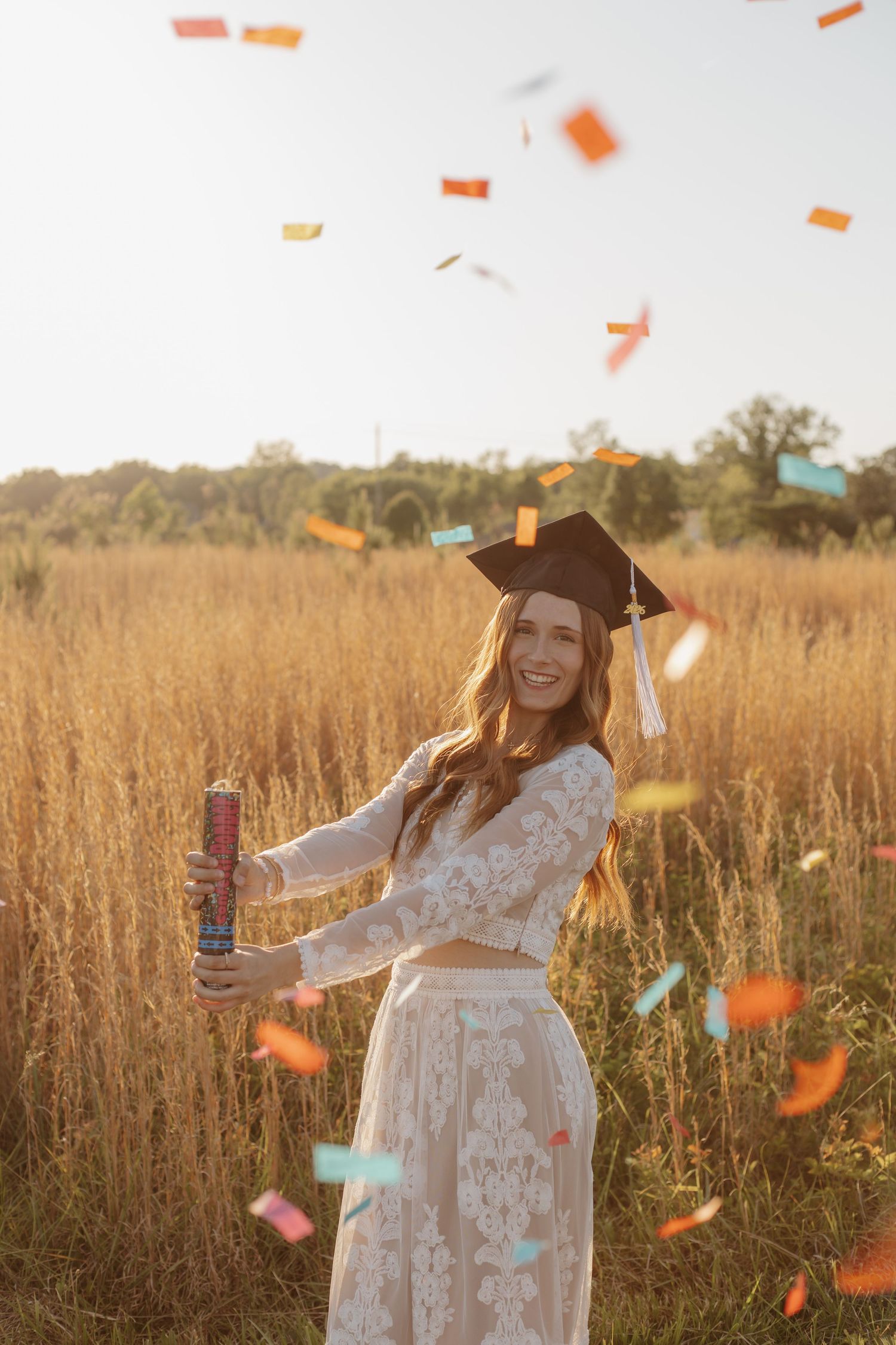 Graduate in cap and white lace dress celebrating with colorful confetti outdoors.