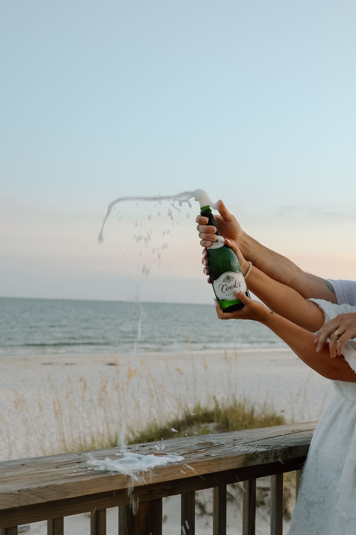 Champagne bottle popping with liquid spraying at beach celebration during golden hour sunset.