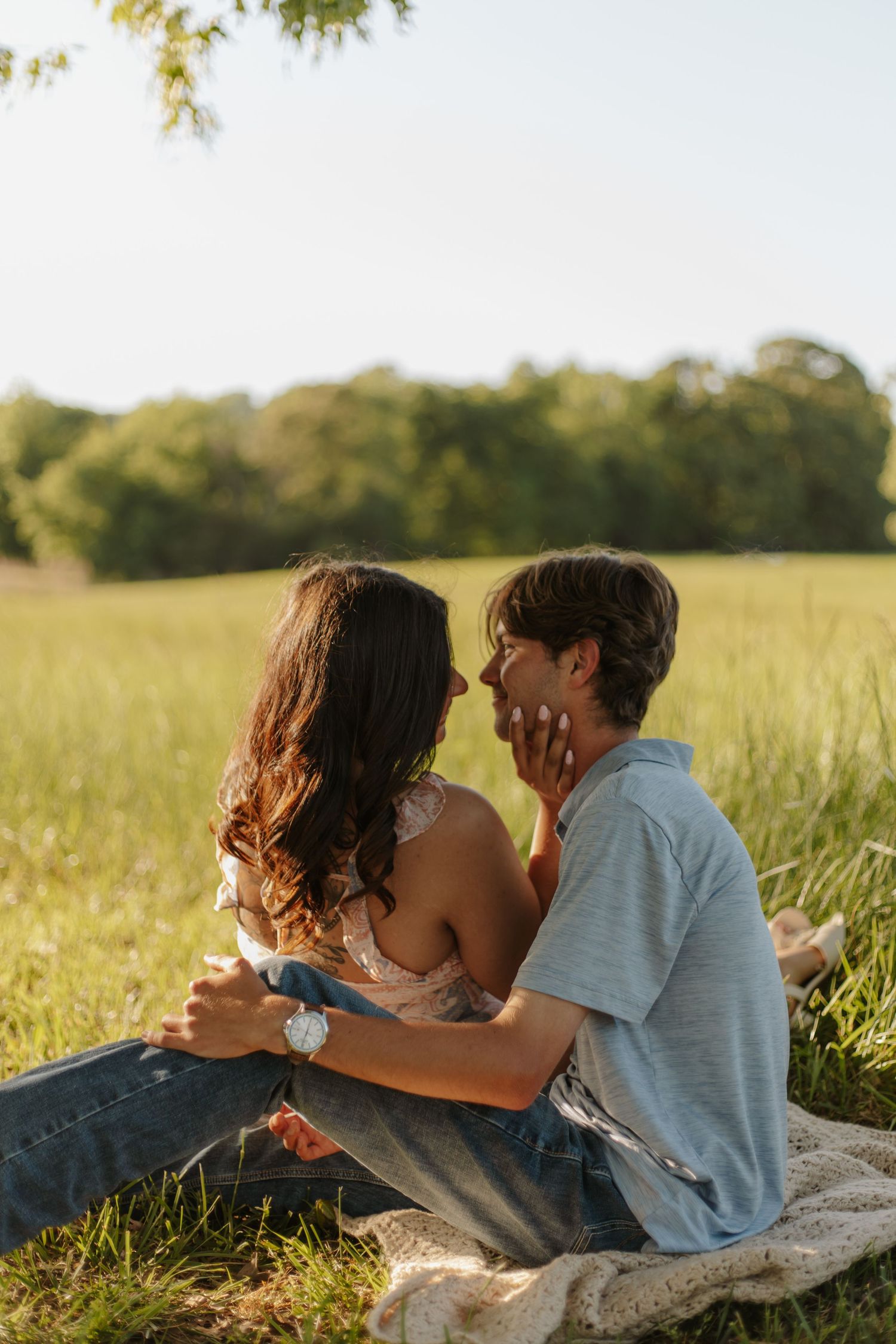 Couple sitting in a sunny green field, sharing a romantic moment as the woman gently touches the man's face.