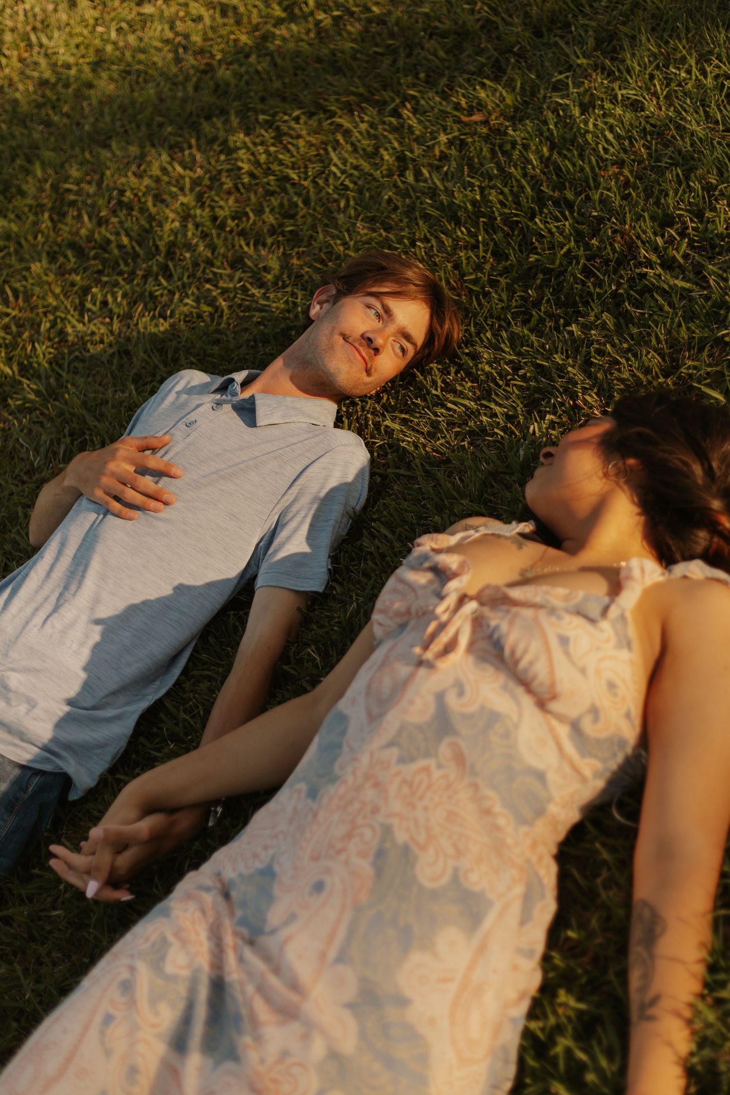 A young couple lies on green grass holding hands in warm golden afternoon sunlight.