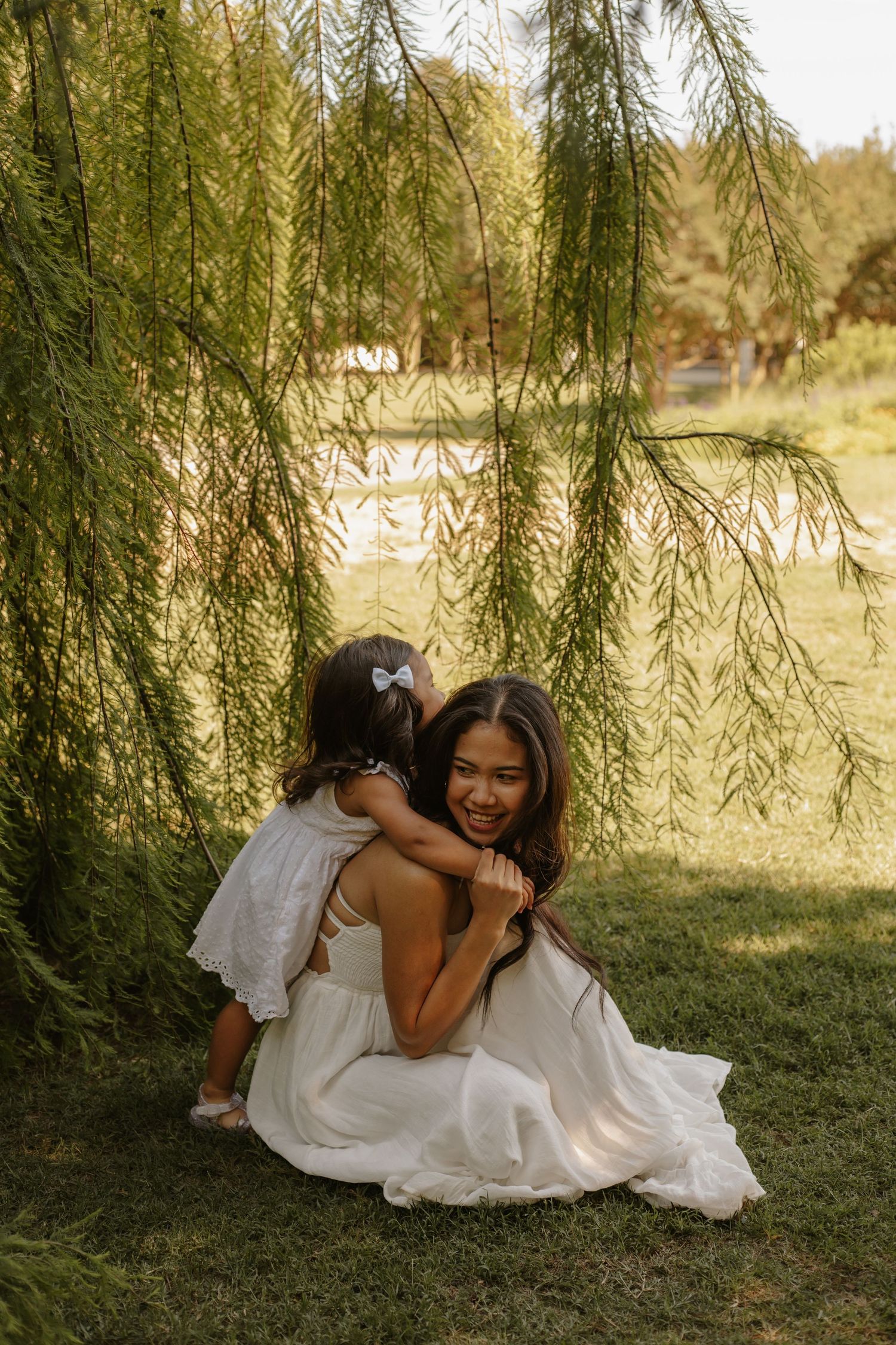 Mother in white dress hugs laughing daughter under weeping willow tree in sunny park.
