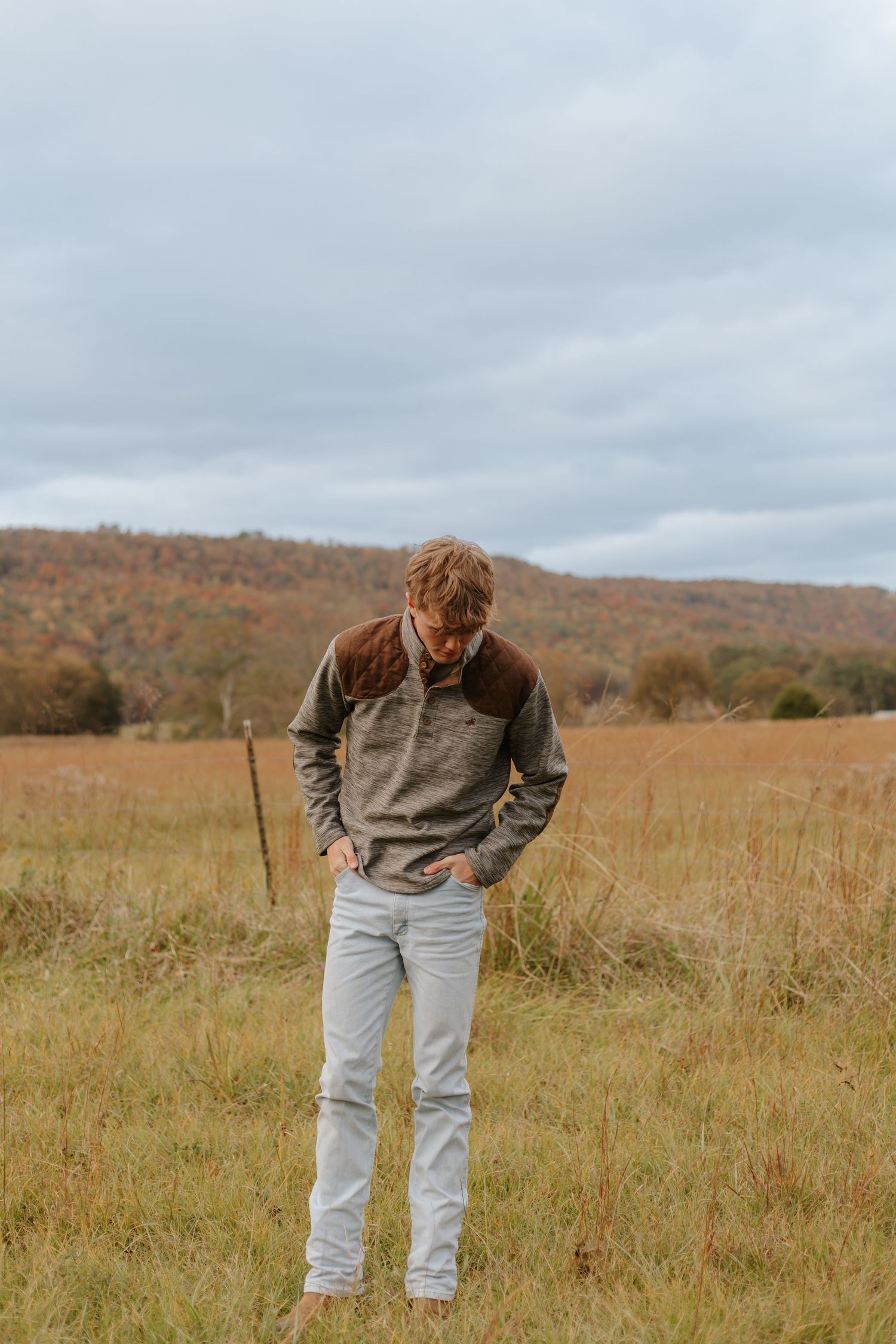 Young man in gray pullover standing in autumn field with mountain backdrop.