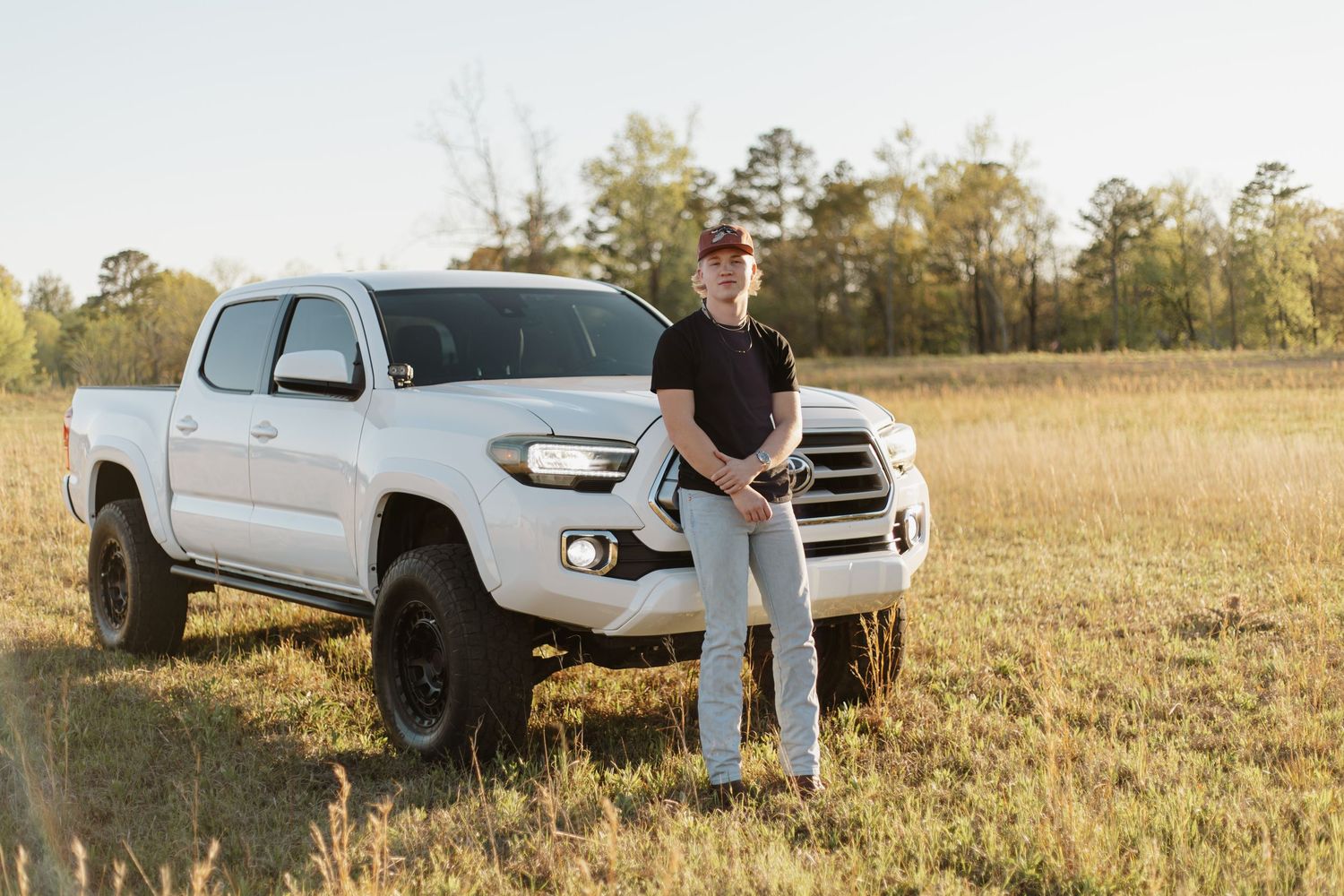 Young man in black shirt leans against white Toyota Tacoma truck in open golden field at sunset.