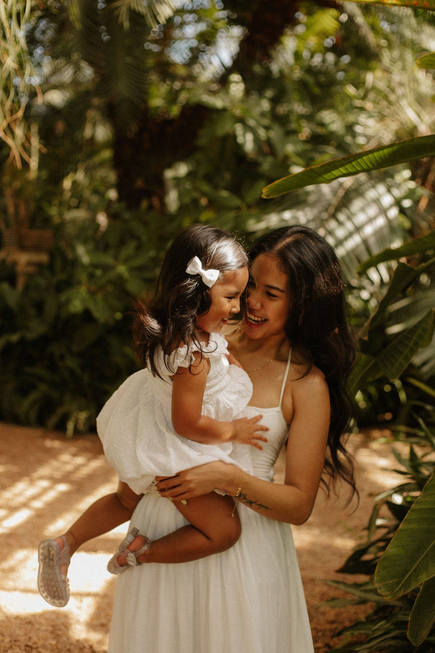 A joyful mother in a white dress holds her laughing toddler daughter in a lush tropical garden setting.