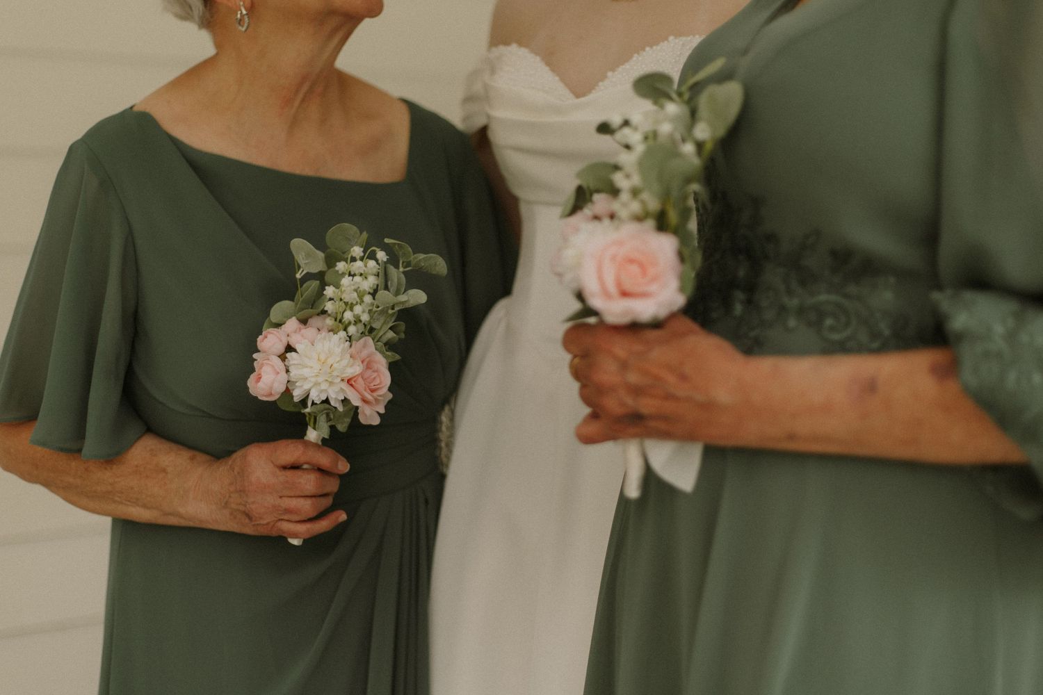 Bride in white gown stands with two women in sage green dresses holding pink floral bouquets at a wedding.