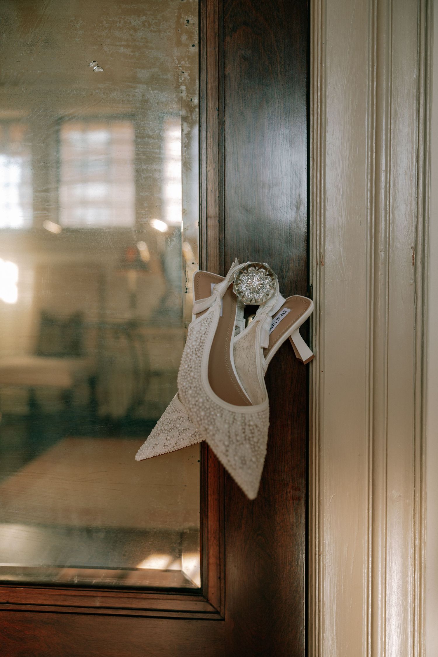 Elegant lace bridal heels with crystal detail hanging on vintage wooden door frame.