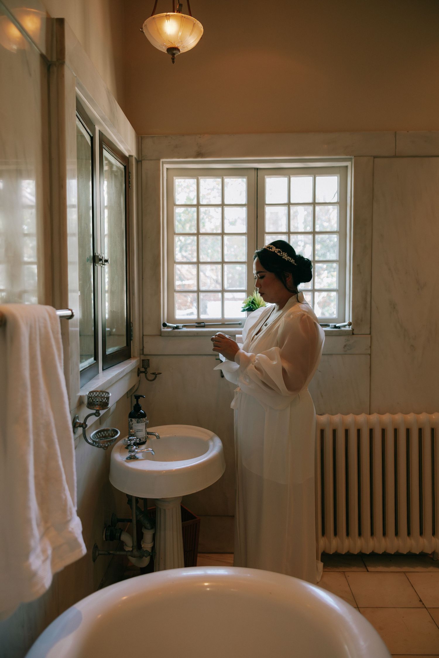 Bride in white robe standing by vintage bathroom window preparing for her wedding day.
