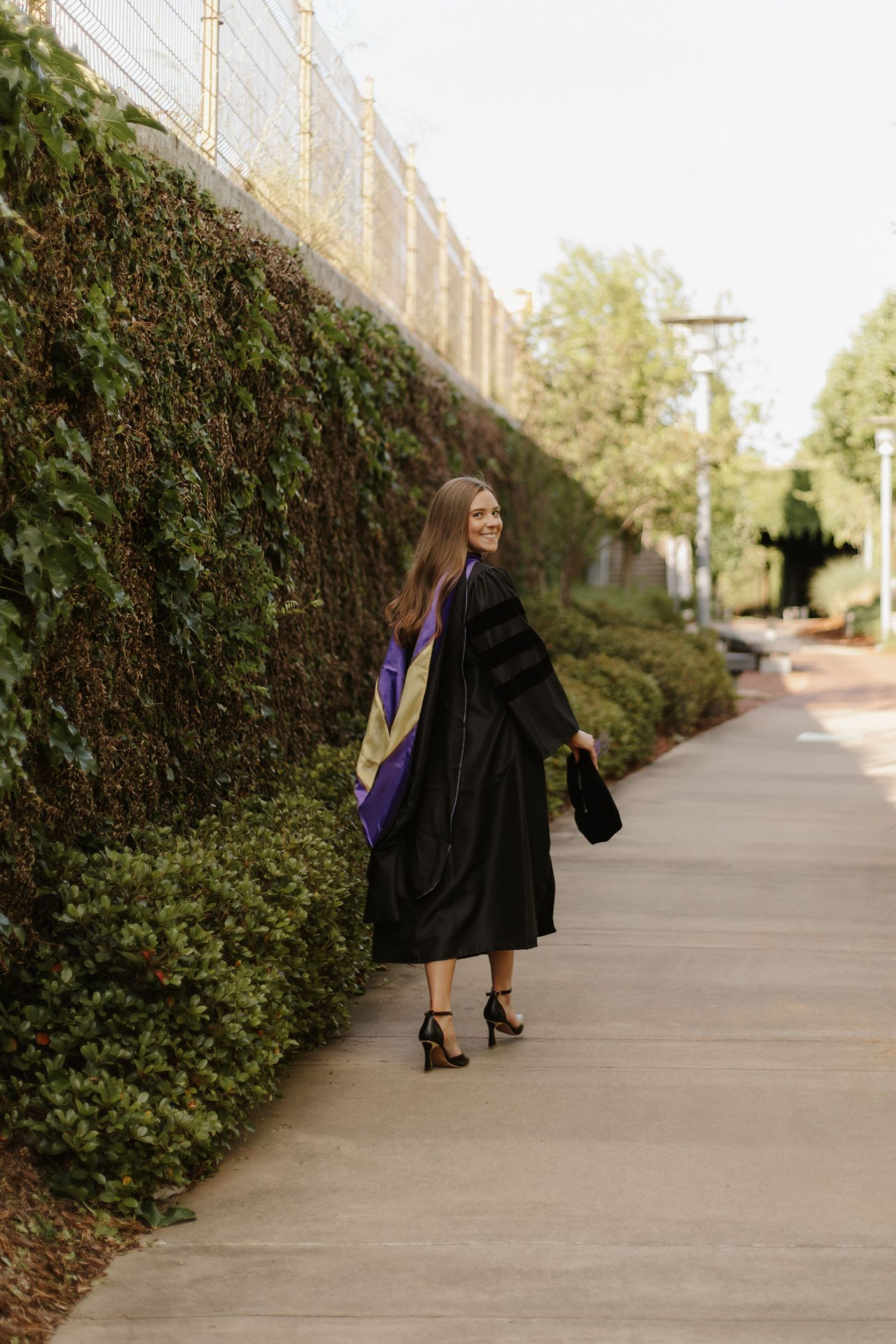 Woman in black graduation gown with purple hood walking along ivy-covered wall pathway.