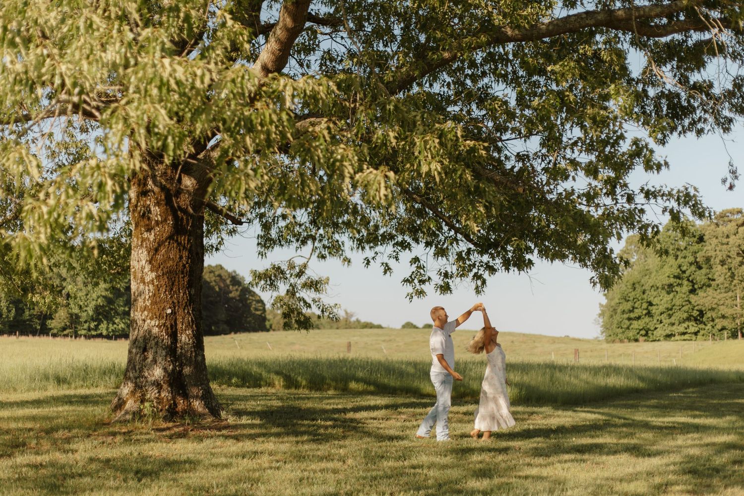 Couple dancing and twirling together under a large tree in a sunlit green meadow.