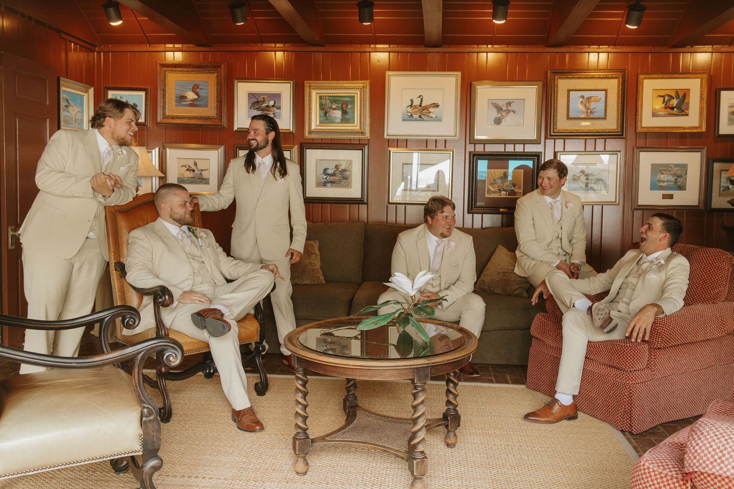 Groomsmen in tan suits relaxing in an elegant wood-paneled room with framed artwork before a wedding ceremony.