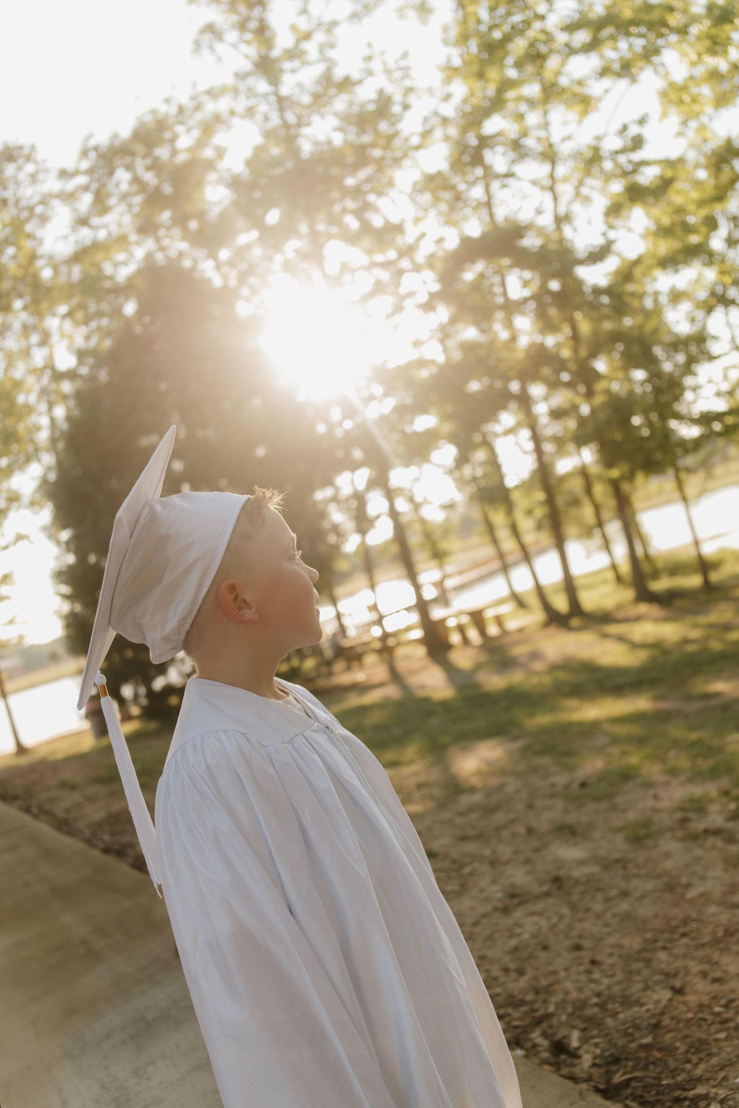 Child in white graduation cap and gown looks up at sunlight filtering through forest trees.