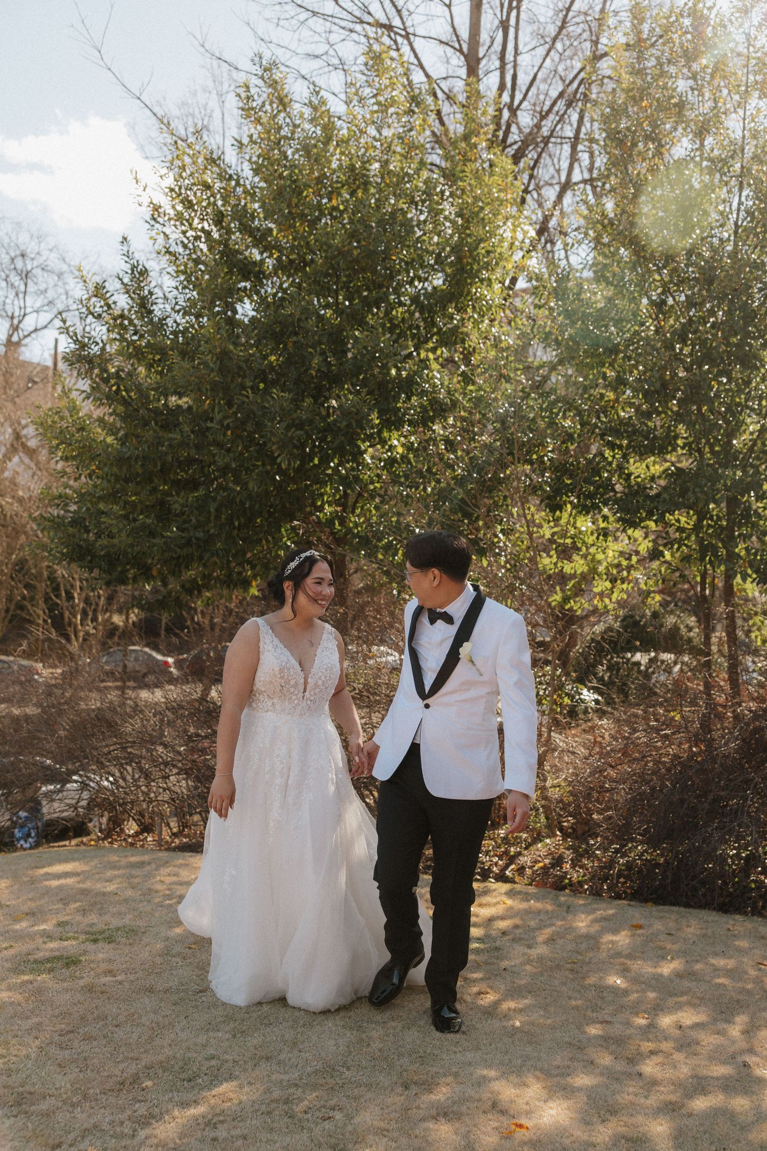 Bride and groom in white tuxedo holding hands walking through a sunlit outdoor garden.