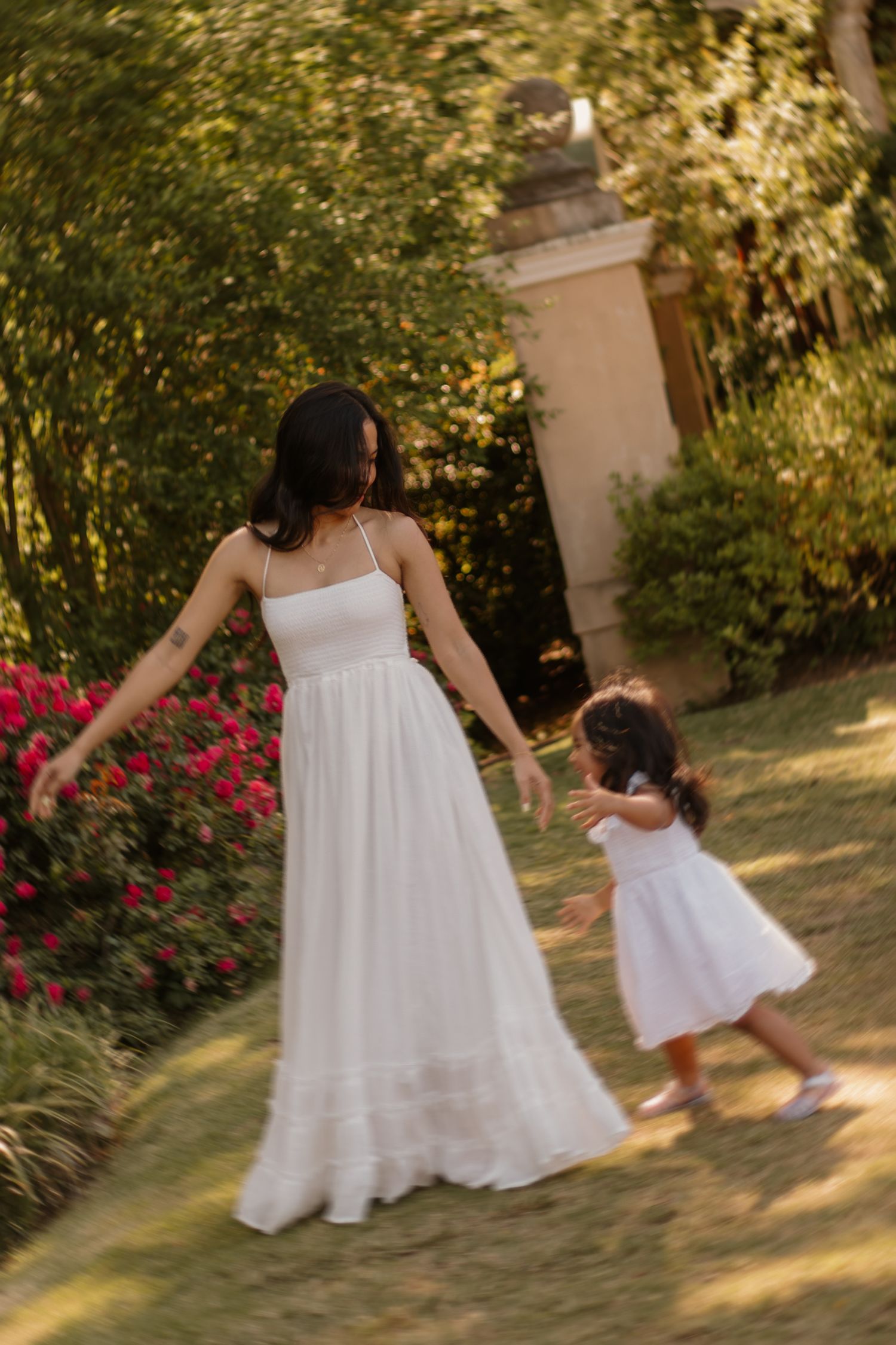 Mother in white maxi dress holds daughters hand walking through lush garden with pink roses.