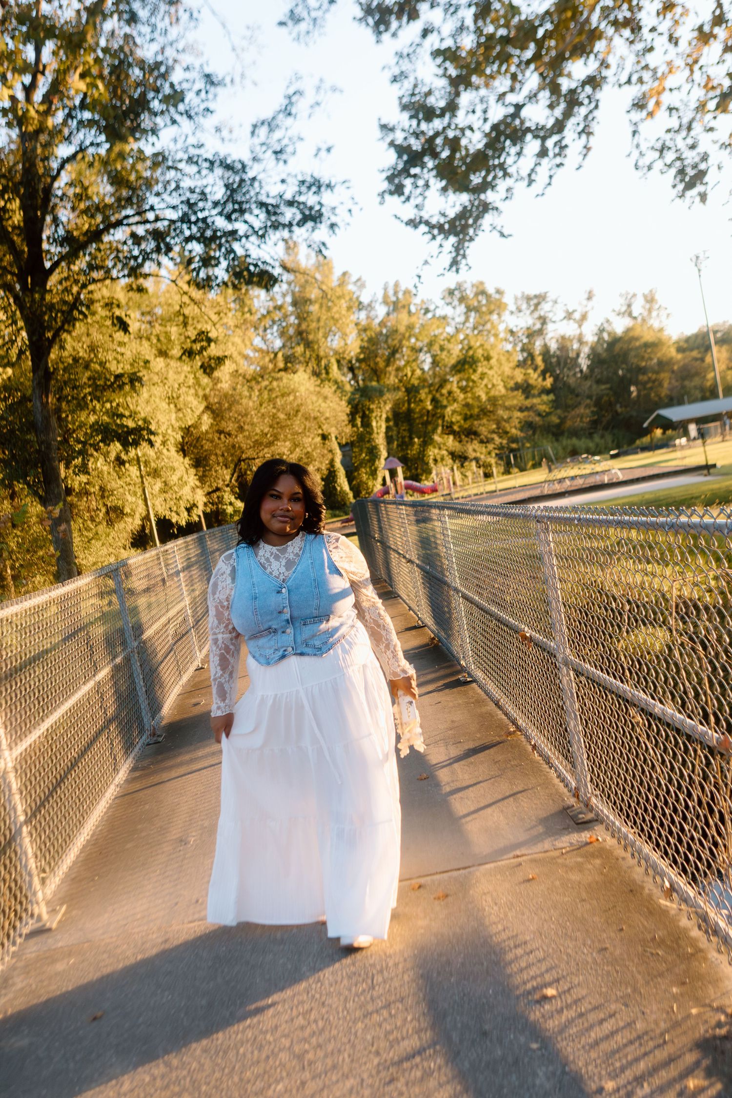 Smiling woman in white skirt and blue vest posing on fenced bridge in golden hour light.