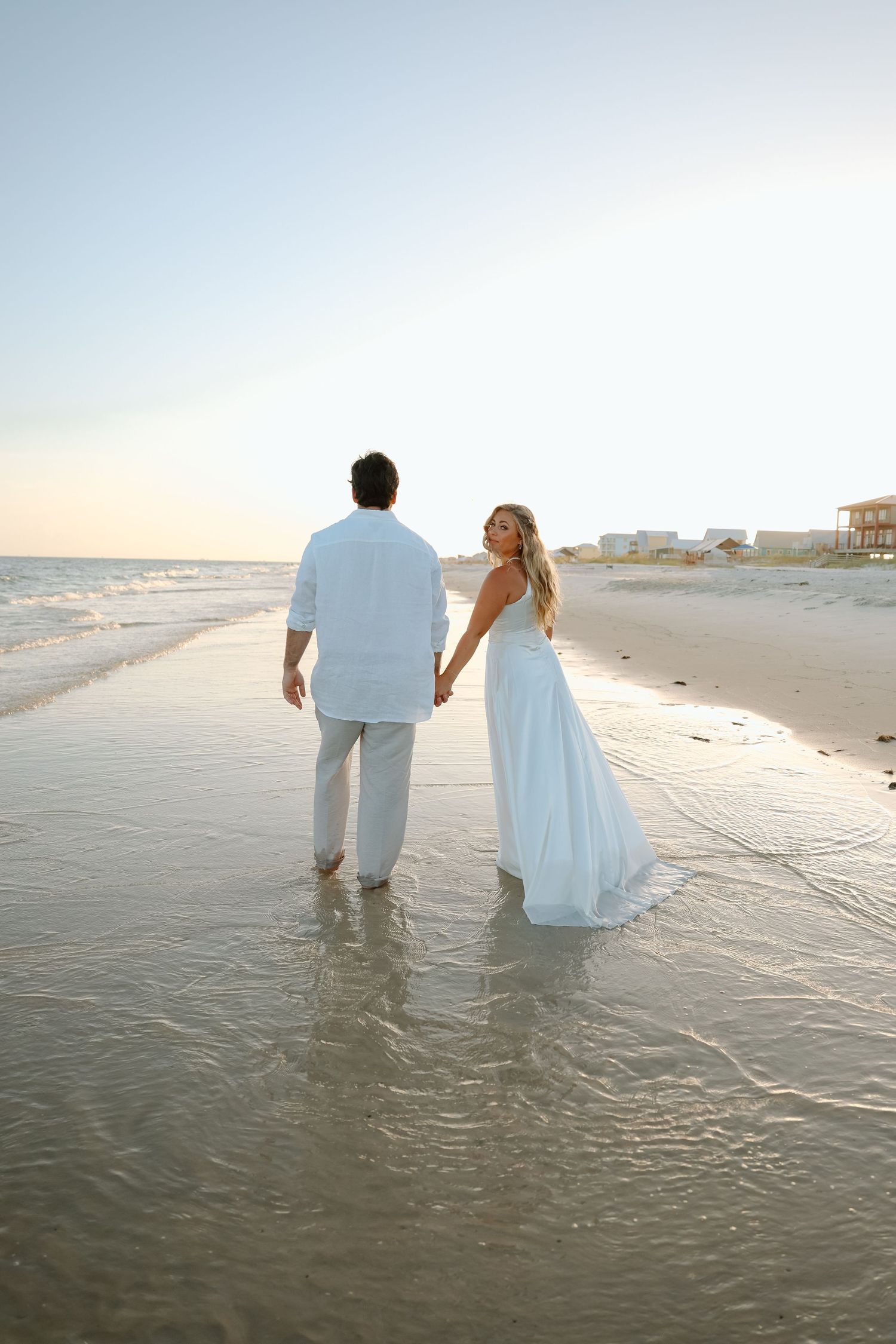 Bride and groom walk hand in hand along shallow ocean shoreline at sunset on their wedding day.