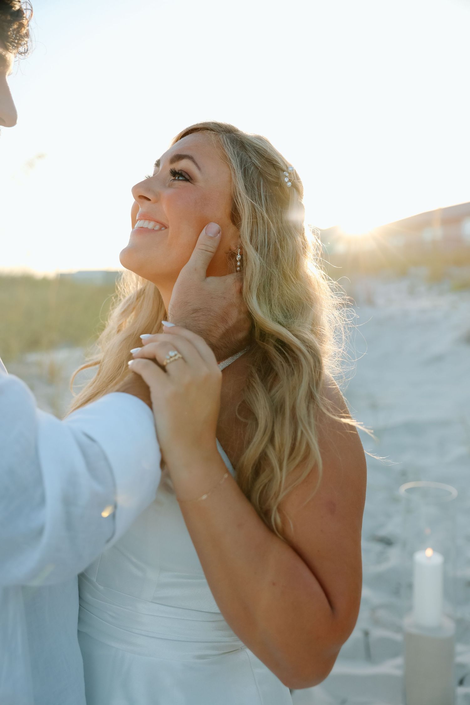 Glowing bride smiles up at groom as he tenderly cups her face during beach wedding ceremony.
