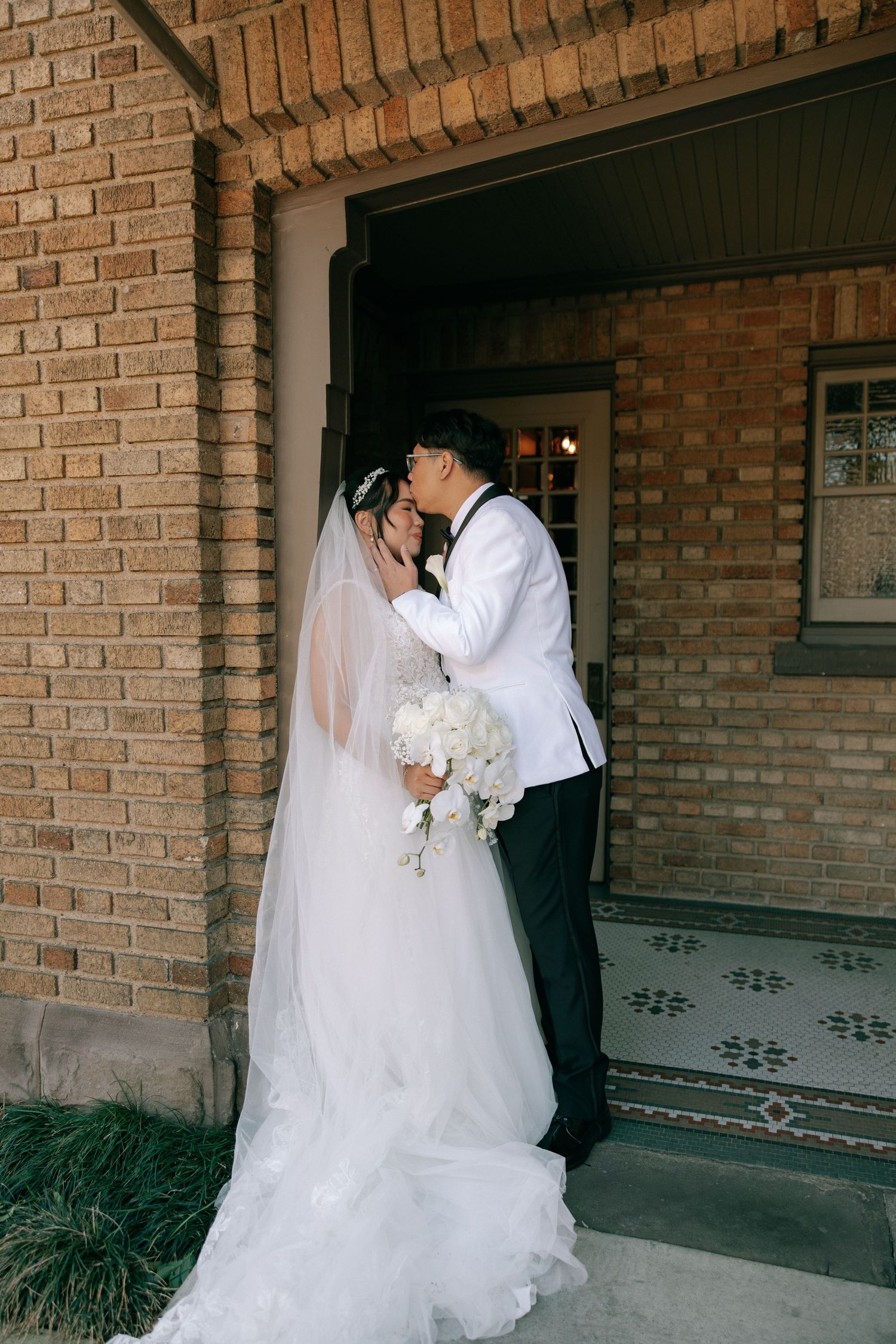 Bride and groom sharing romantic kiss at brick building entrance, cathedral veil trailing behind bride.