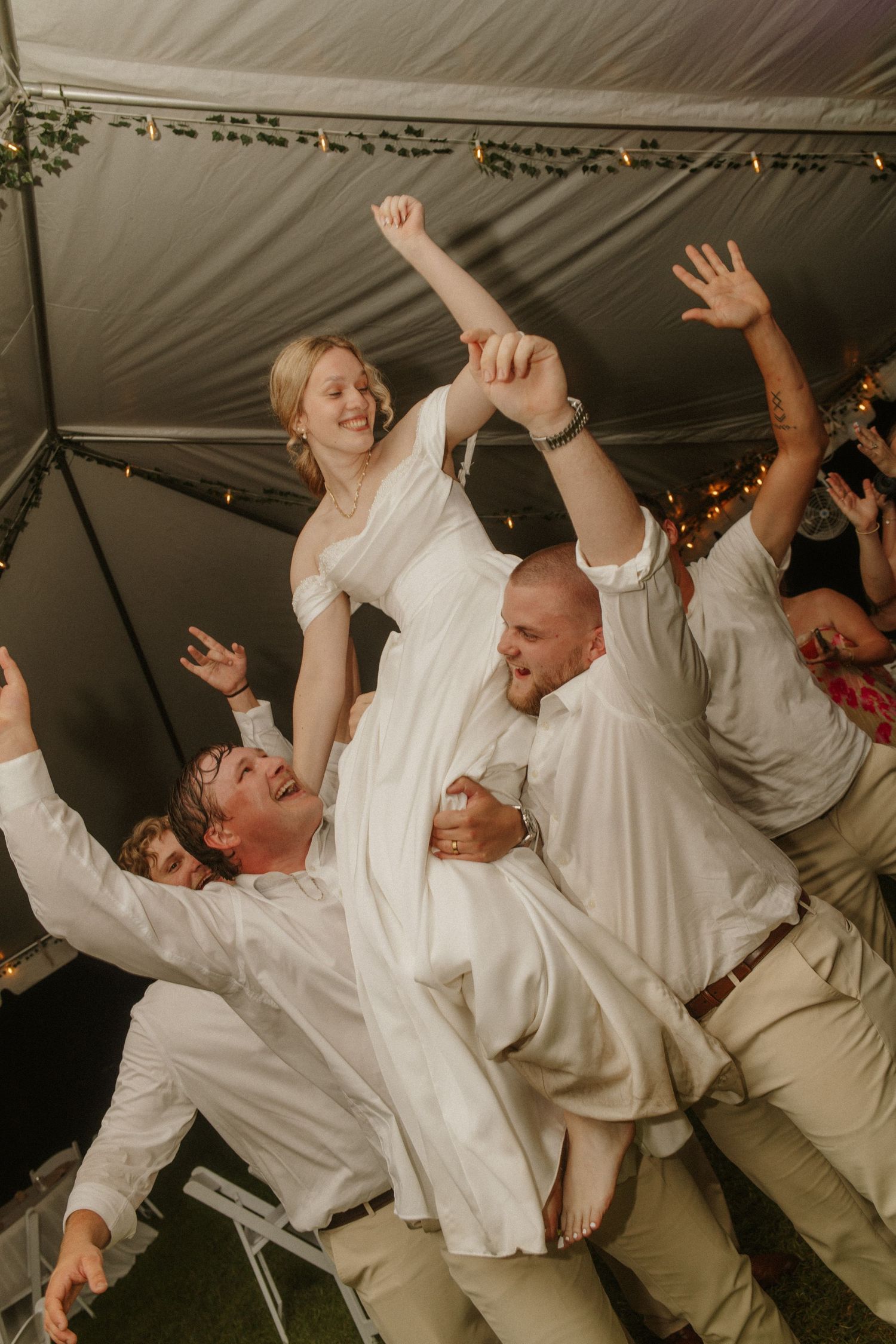 Bride in white dress lifted by groomsmen celebrating joyfully under a tent at a lively wedding reception.