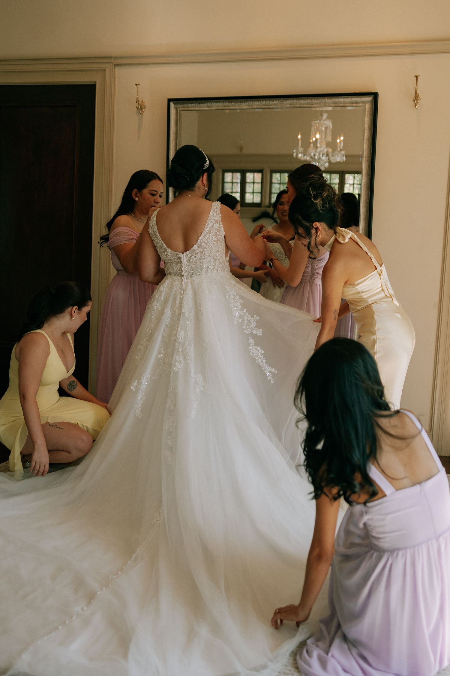 Bridesmaids in pastel dresses help bride into lace wedding gown before mirror in bridal suite.
