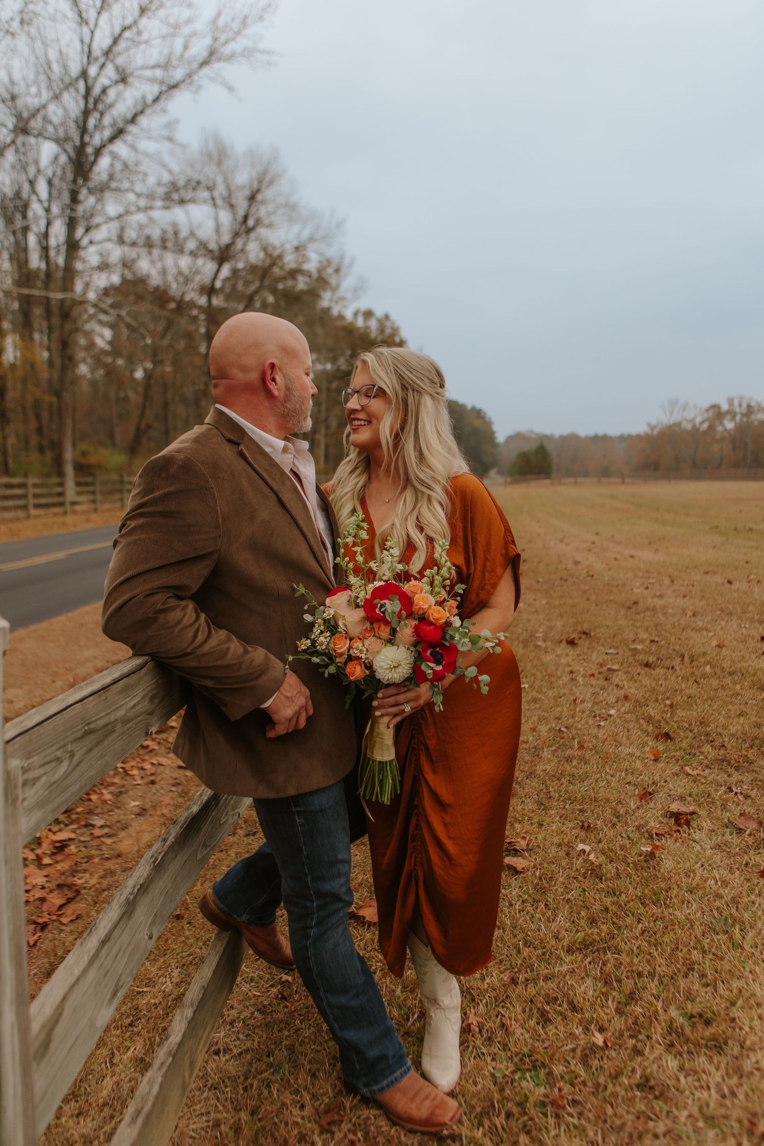 Couple leaning against wooden fence in autumn field, bride in rust dress holding colorful bridal bouquet.
