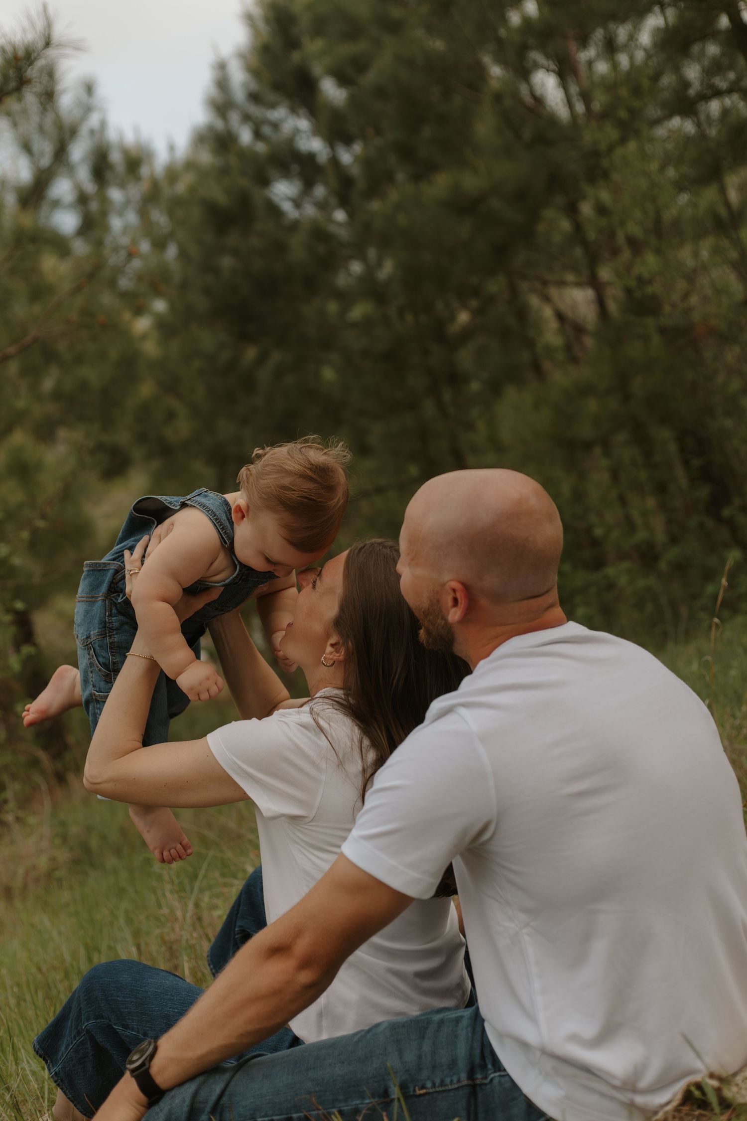 Parents in white shirts sit in meadow lifting toddler in denim overalls for a kiss.