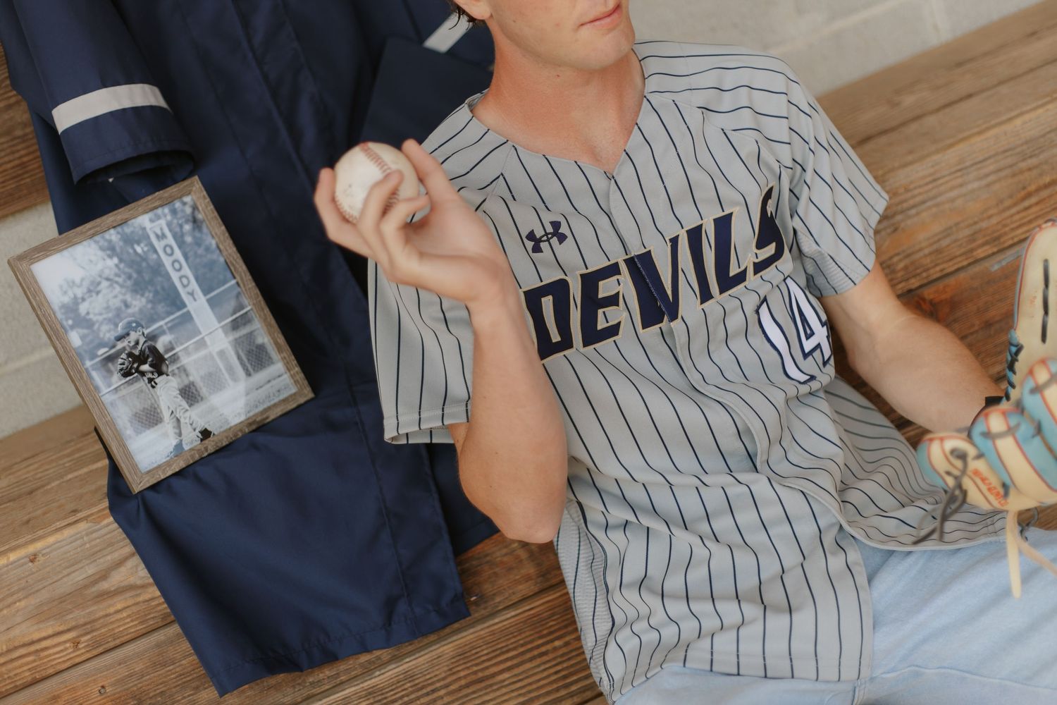 High school senior in Devils baseball jersey holds a ball beside a framed photo and uniform.