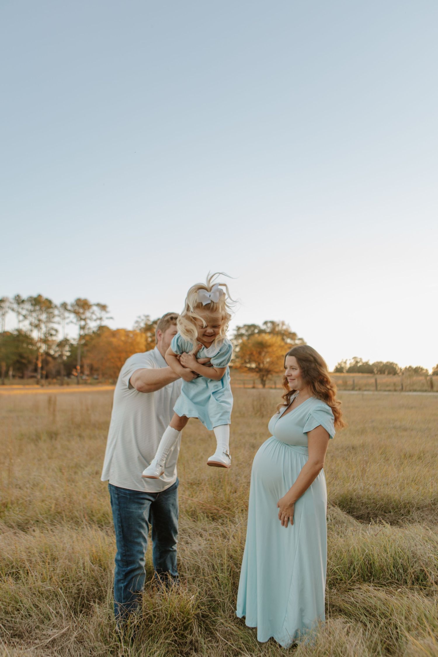Pregnant woman in mint dress watches husband lift toddler in sunlit golden field at dusk.