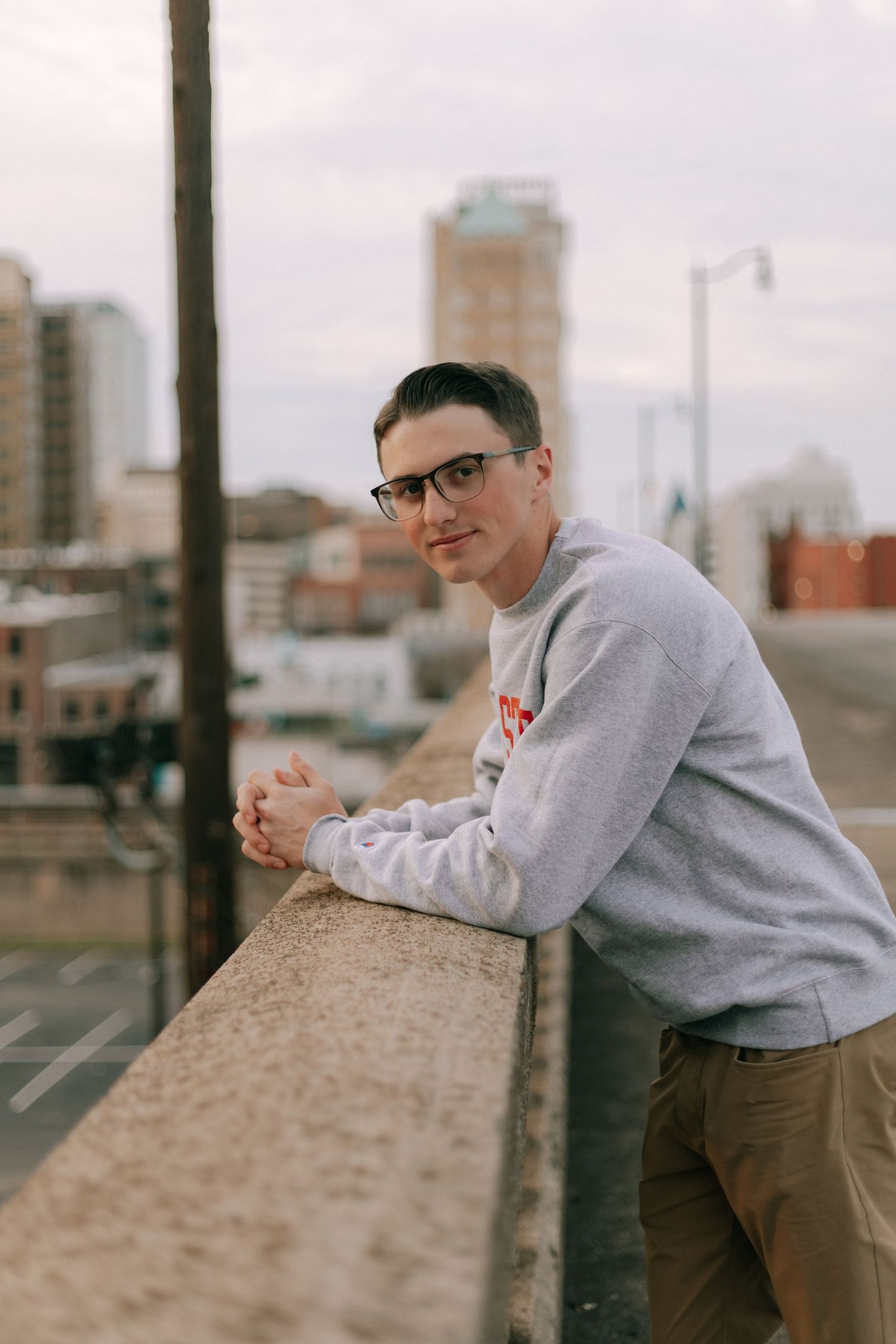 Young man in gray sweatshirt leaning on urban rooftop railing with city skyline background.