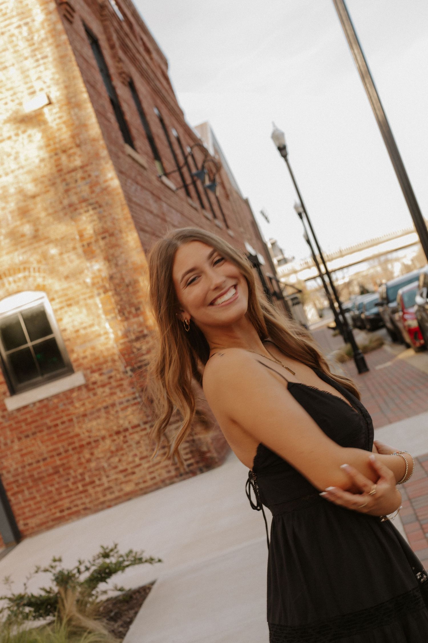 Smiling young woman in black dress poses on urban sidewalk with brick building background.