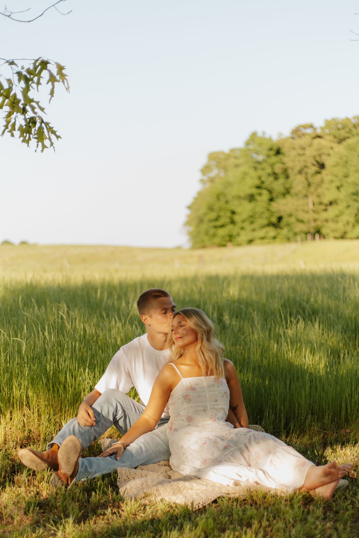 Couple sitting together in a sunny green field, man kissing womans cheek as she smiles softly.