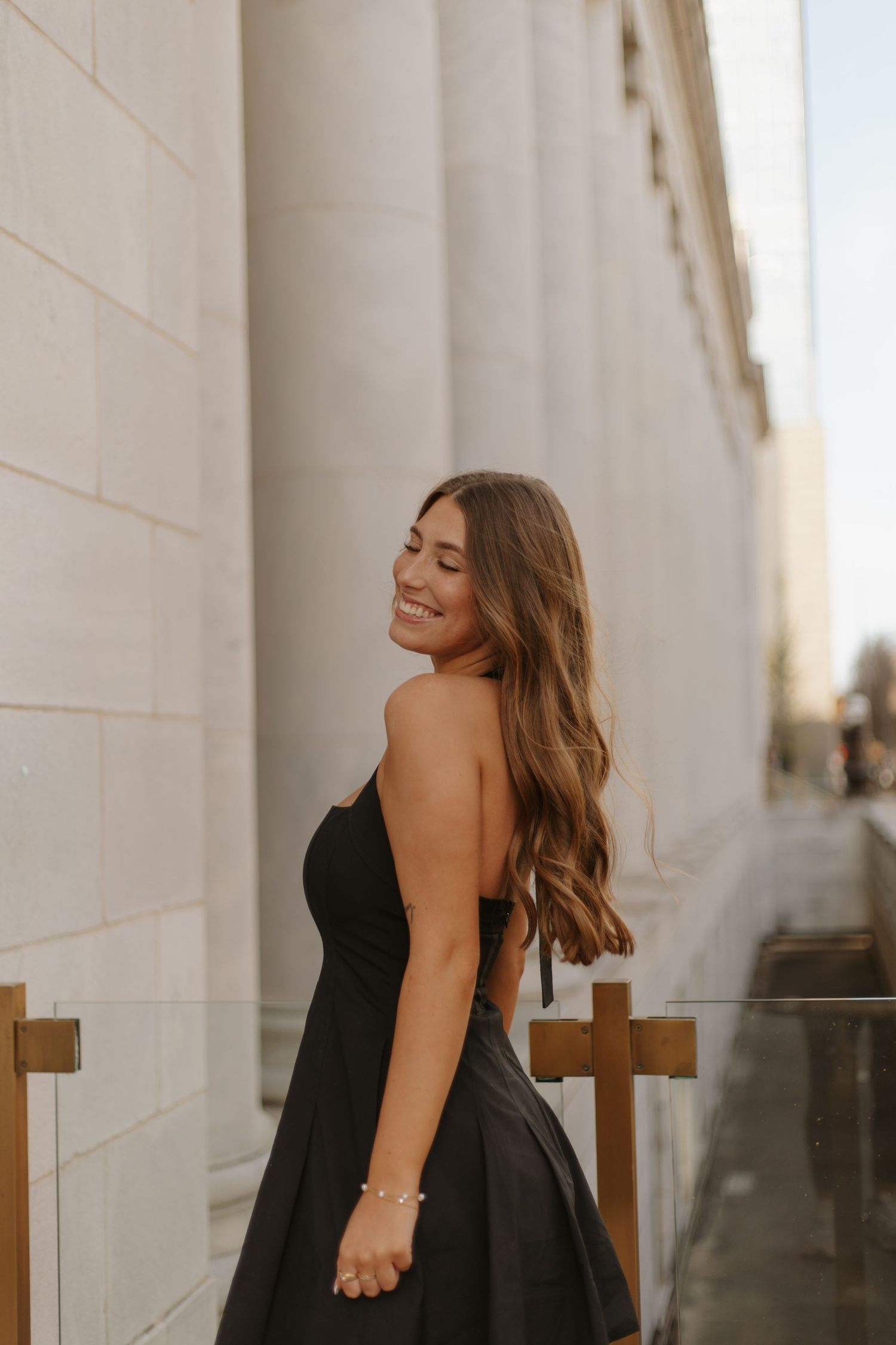 Smiling brunette woman in black strapless dress poses beside white stone columns at golden hour.