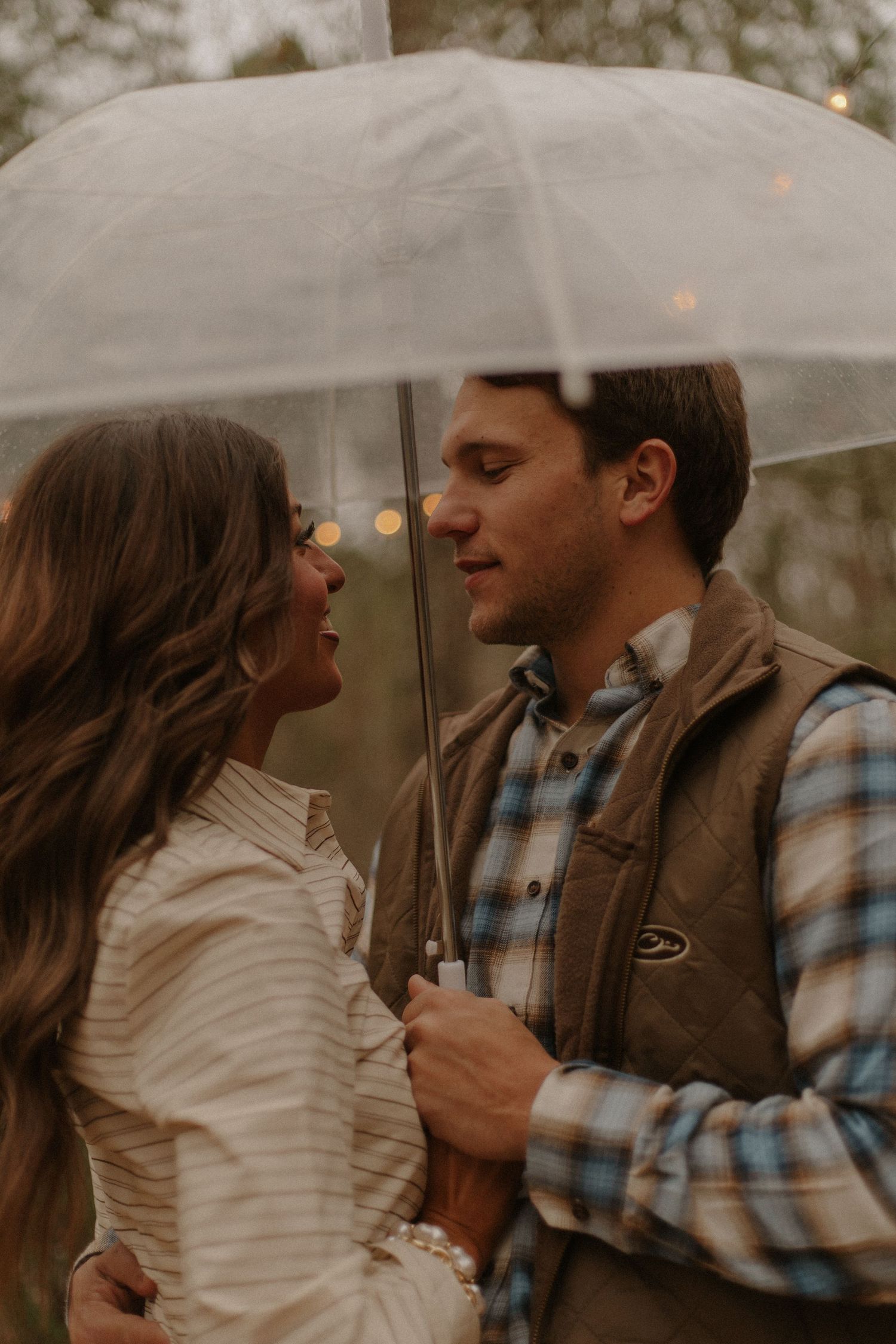 Romantic couple sharing moment under clear umbrella in rainy outdoor setting with bokeh lights.