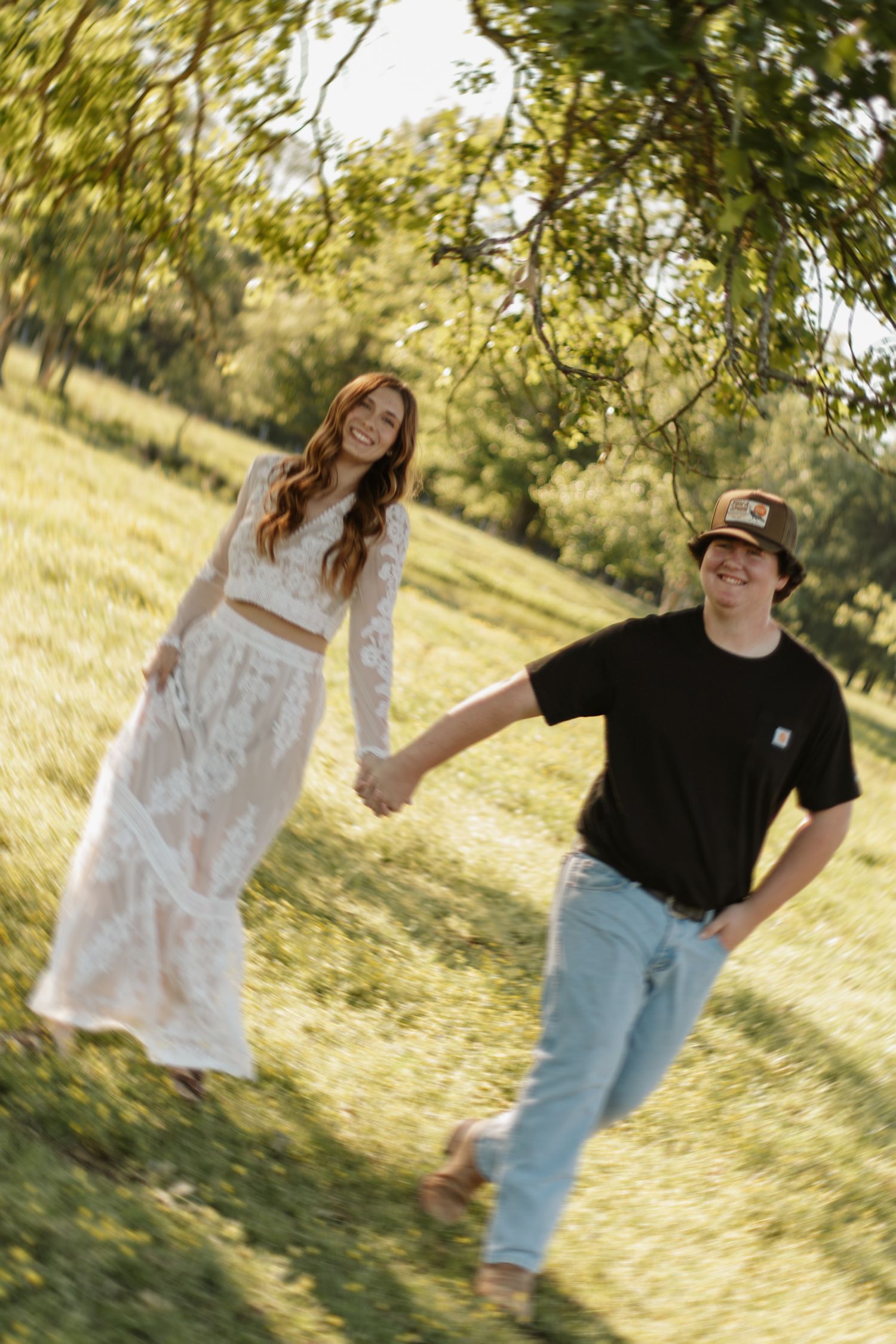 A happy couple holds hands walking through a sunny green park, the woman in a white lace outfit.