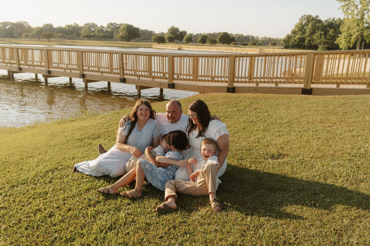Family of five sitting together on green grass by a wooden dock and serene lake during golden hour.