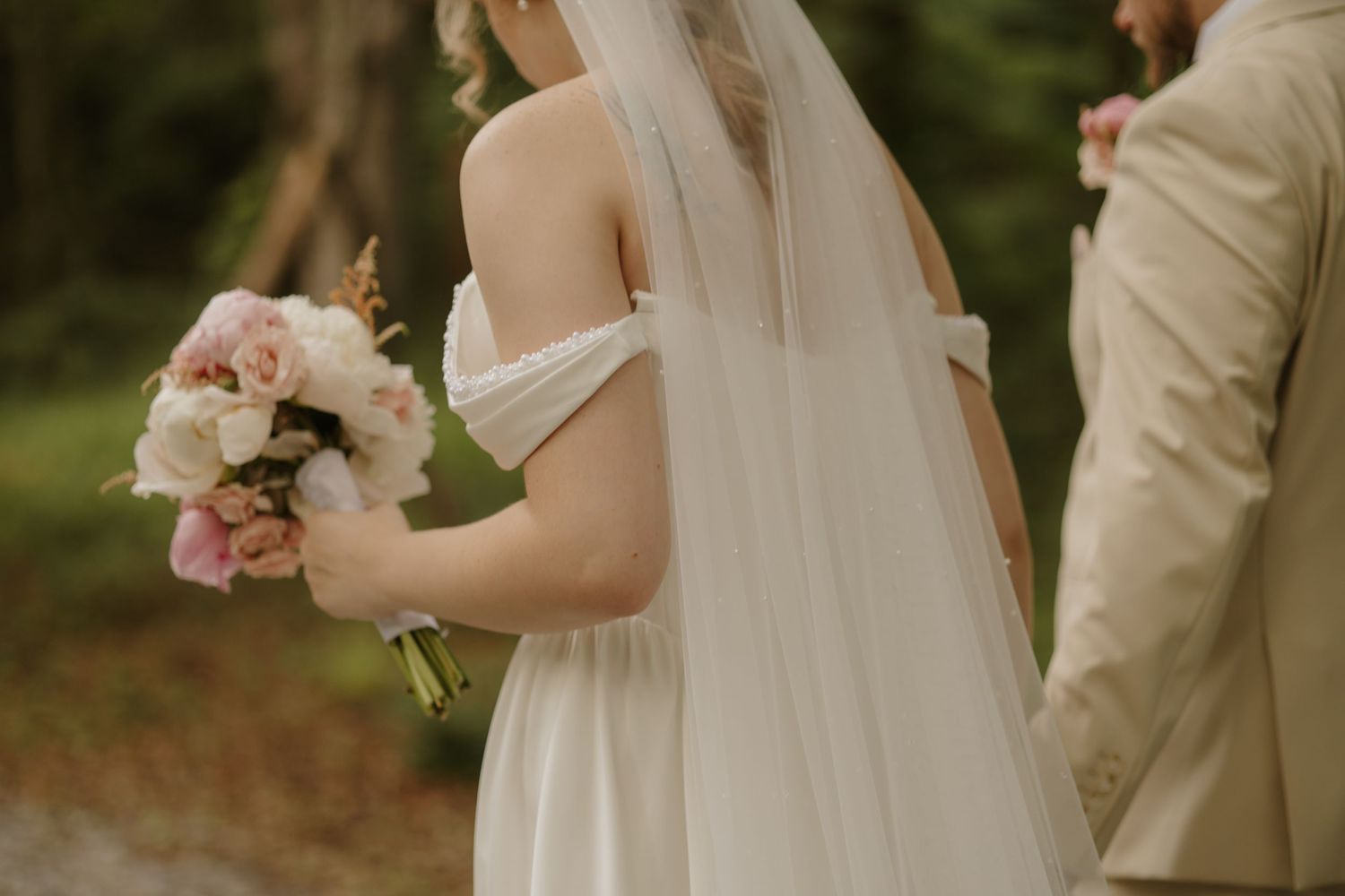 Bride in off-shoulder white gown holding pink and white bouquet, standing beside groom in tan suit outdoors.