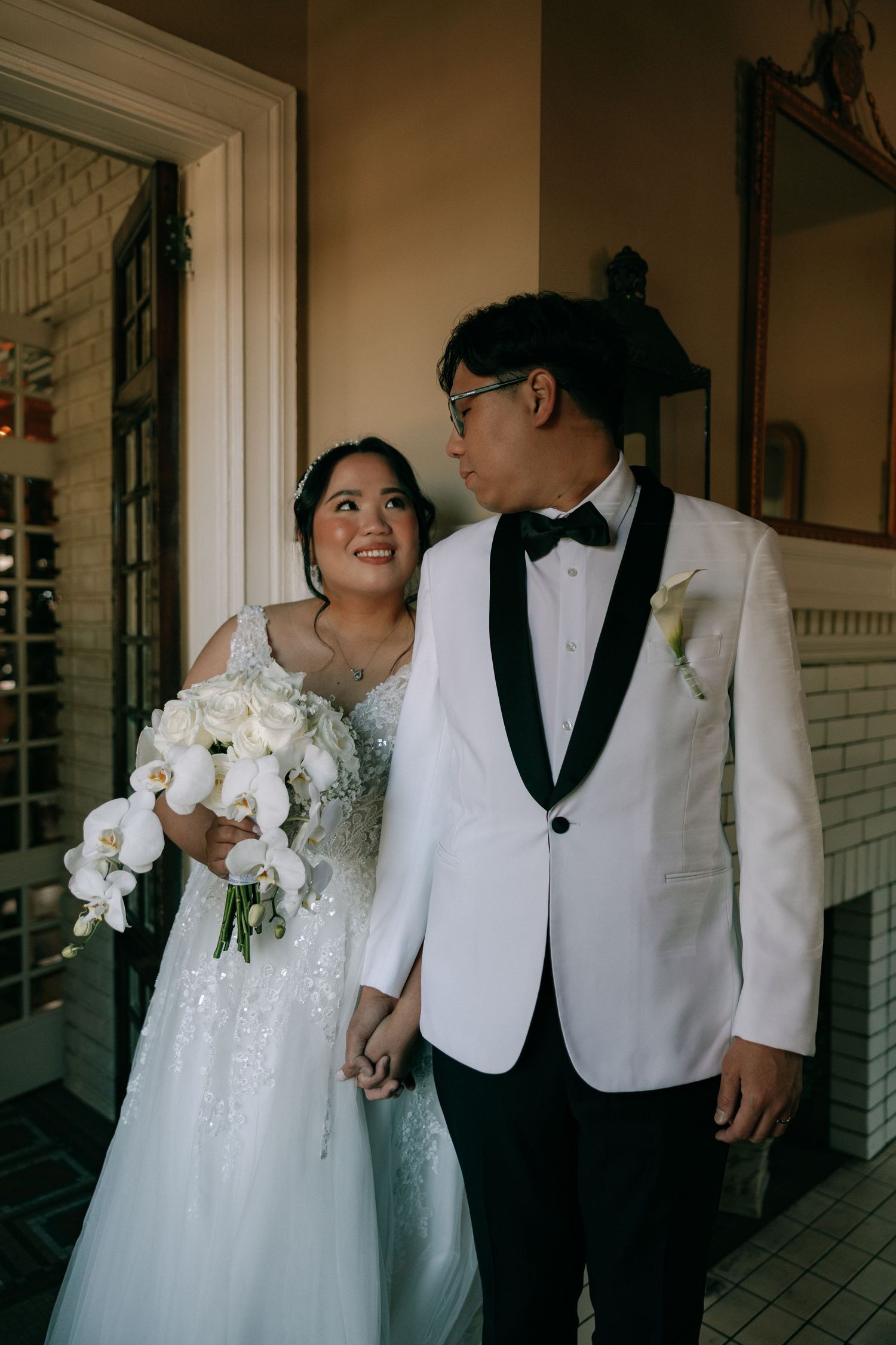 Bride and groom holding hands indoors, bride carrying white orchid bouquet in elegant lace wedding gown.