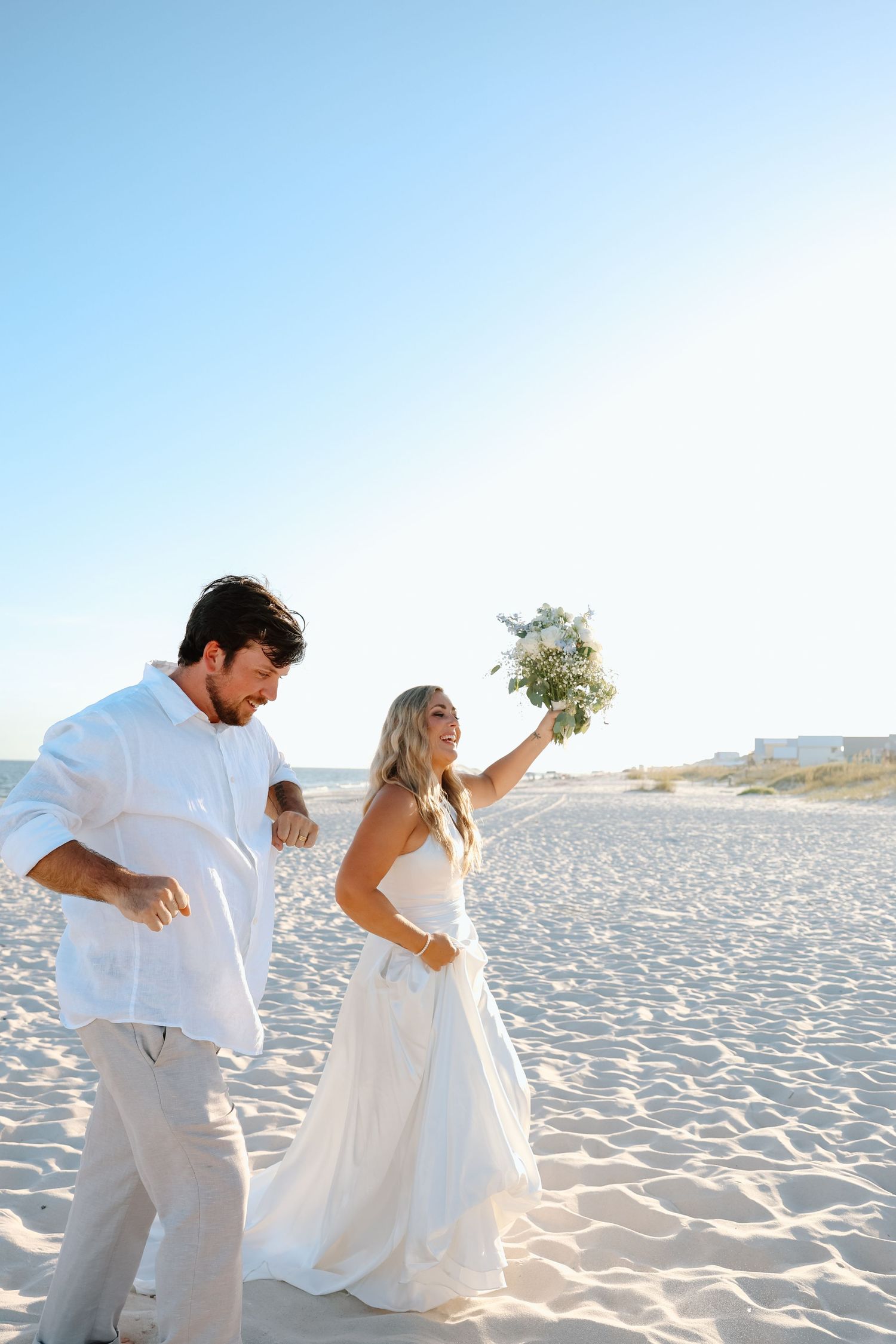 Joyful bride in white gown raises bouquet while groom in white linen walks together on sunny beach.