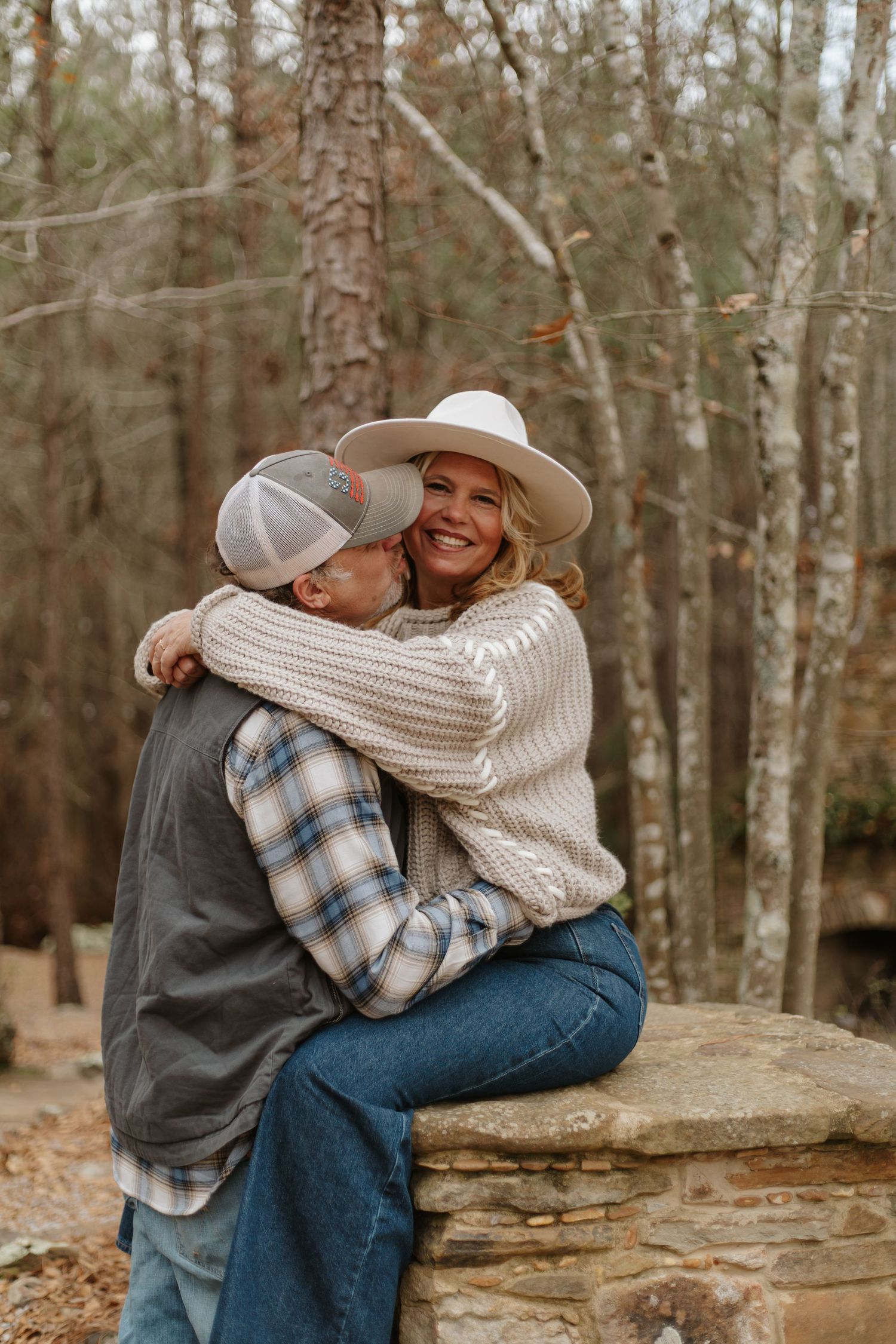 Happy couple embracing on stone wall in forest, woman in white hat and chunky knit sweater.