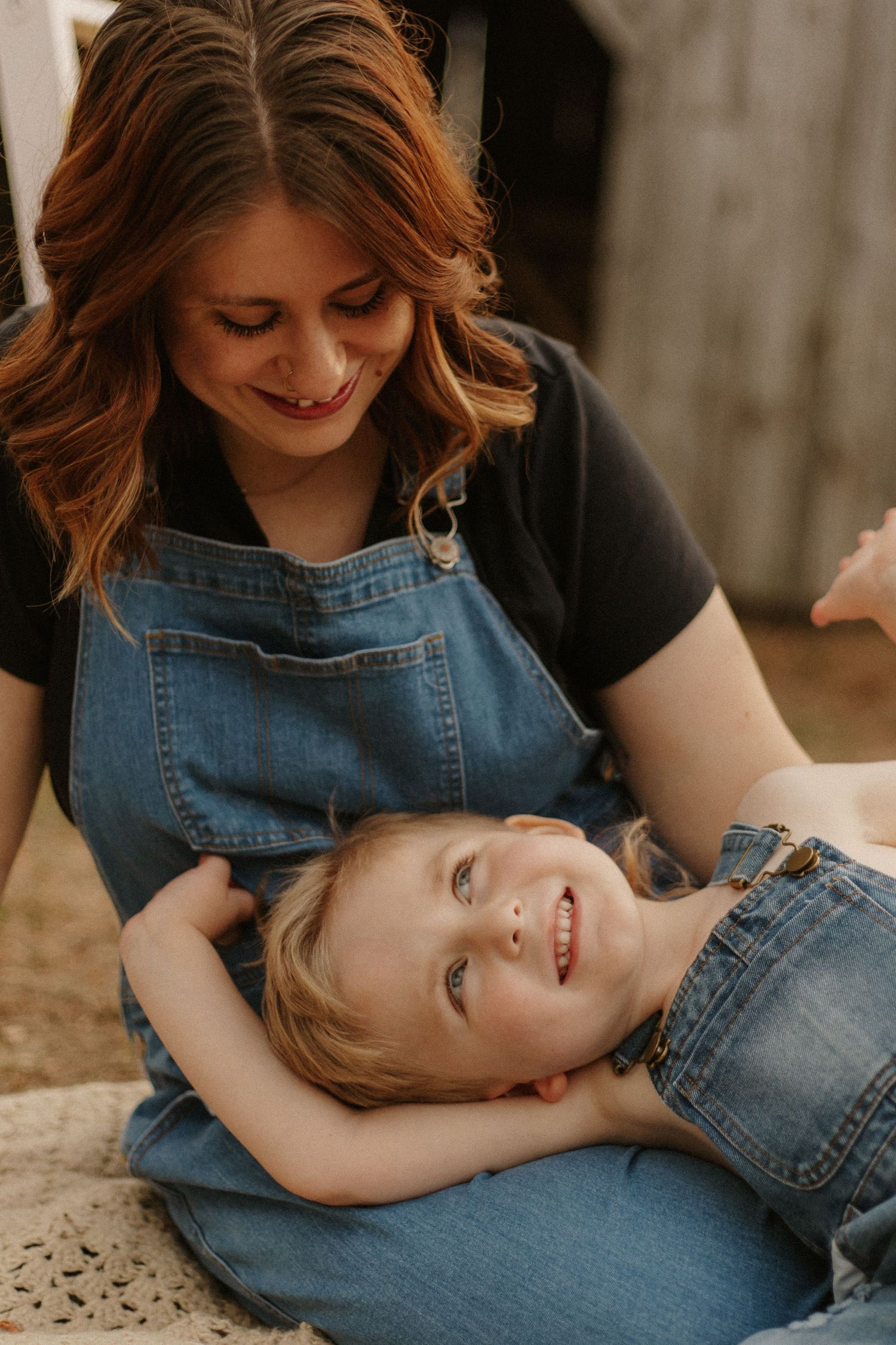 Red-haired mother in black top smiles at blonde child lying in her lap wearing denim overalls.