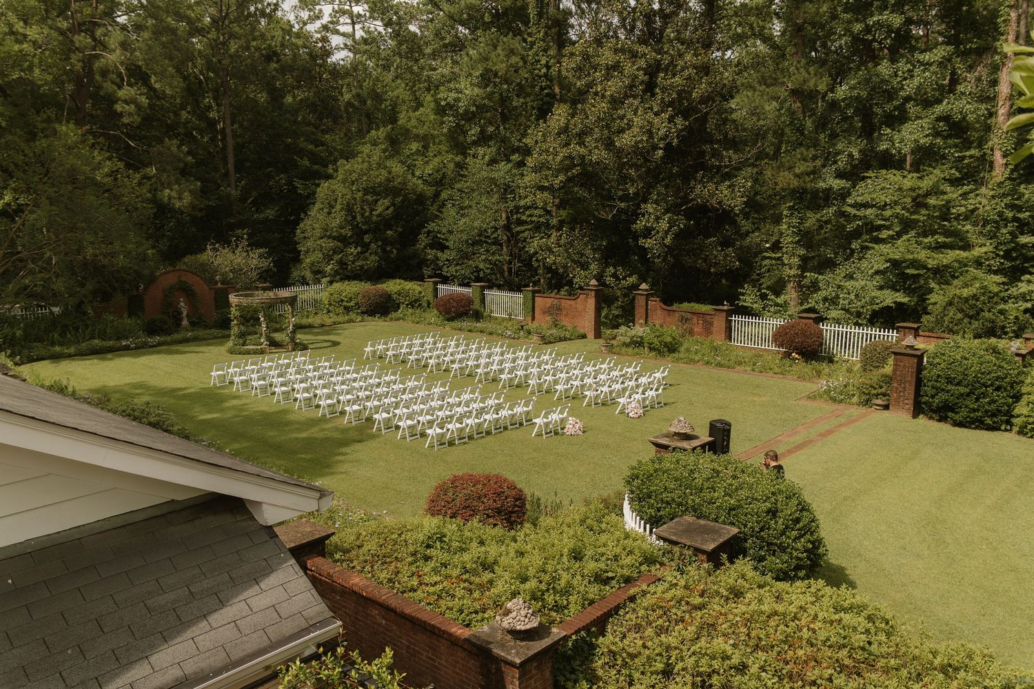 Aerial view of an elegant outdoor wedding ceremony setup with white chairs on a lush green lawn surrounded by trees.