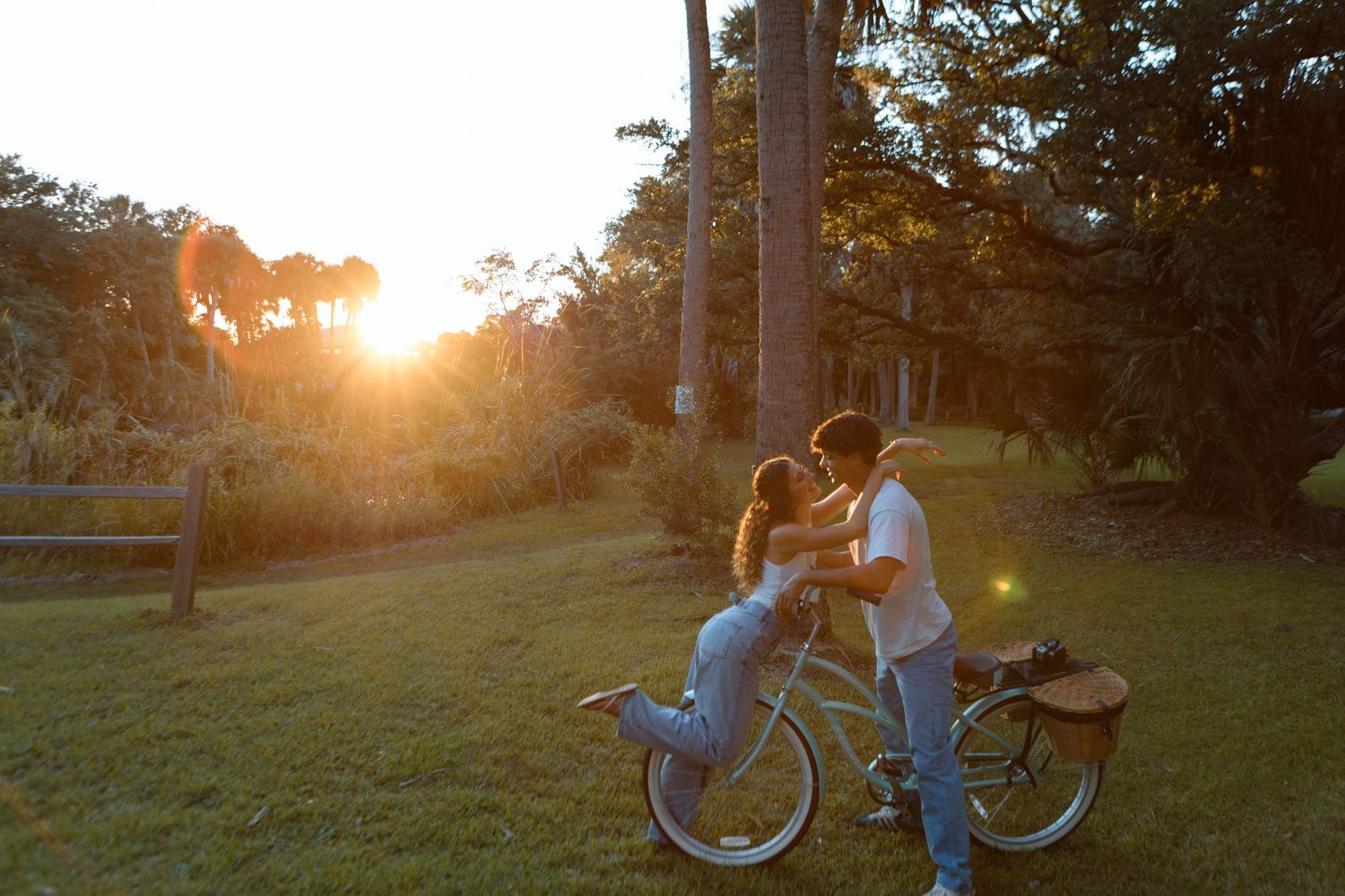 Couple kissing at sunset beside a vintage bicycle in a lush green park with tall trees.