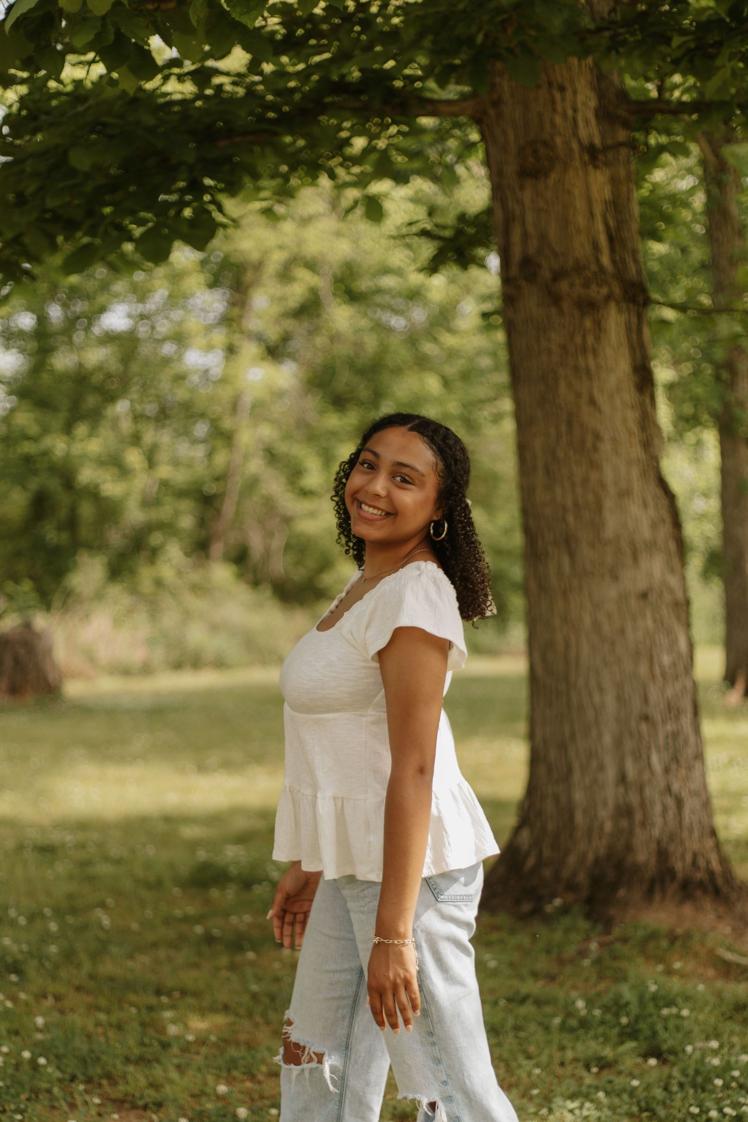 Smiling young woman in white top and ripped jeans posing outdoors near a large tree.