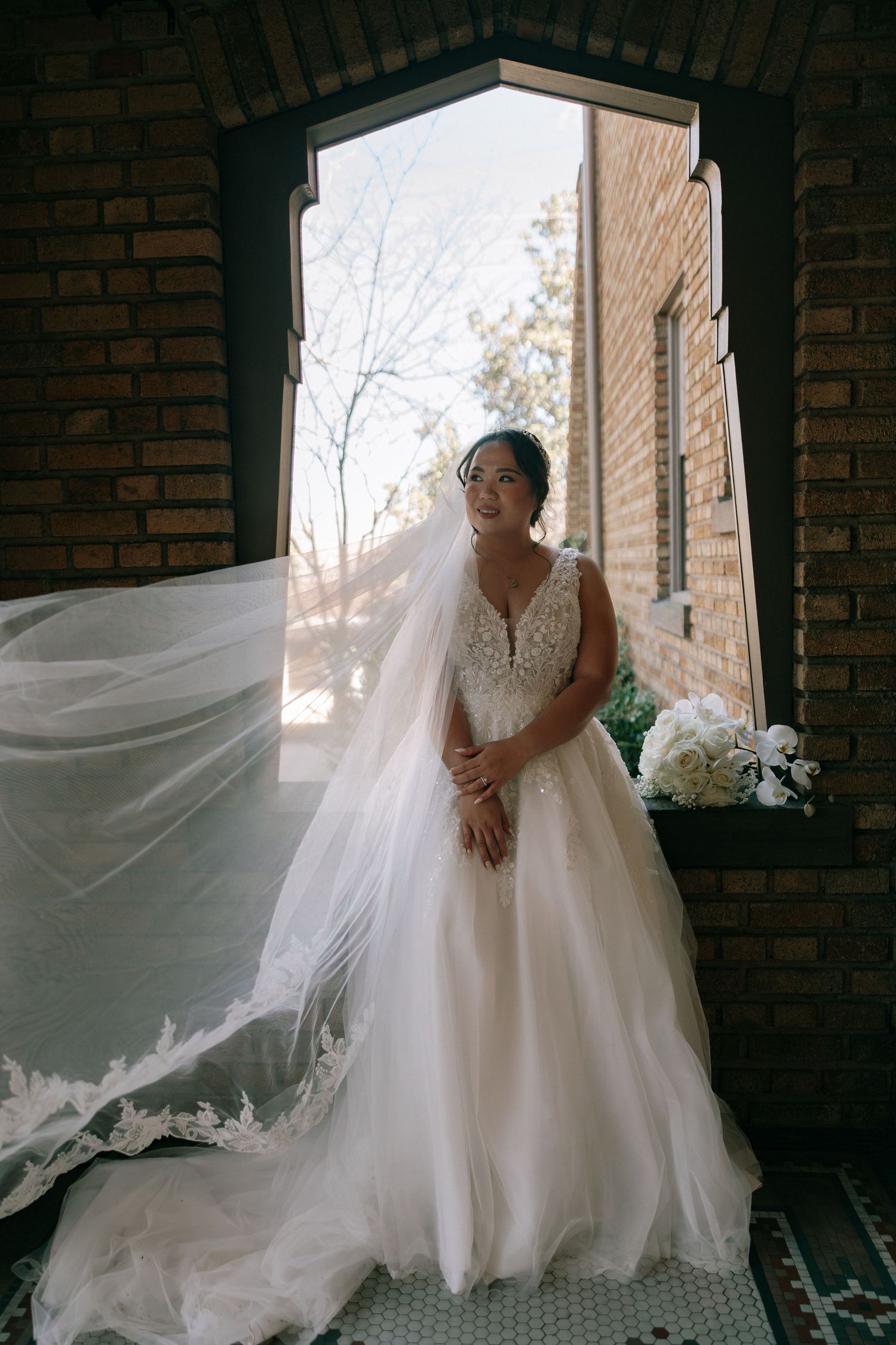 Bride in lace ball gown with flowing cathedral veil standing in arched brick doorway with bouquet.