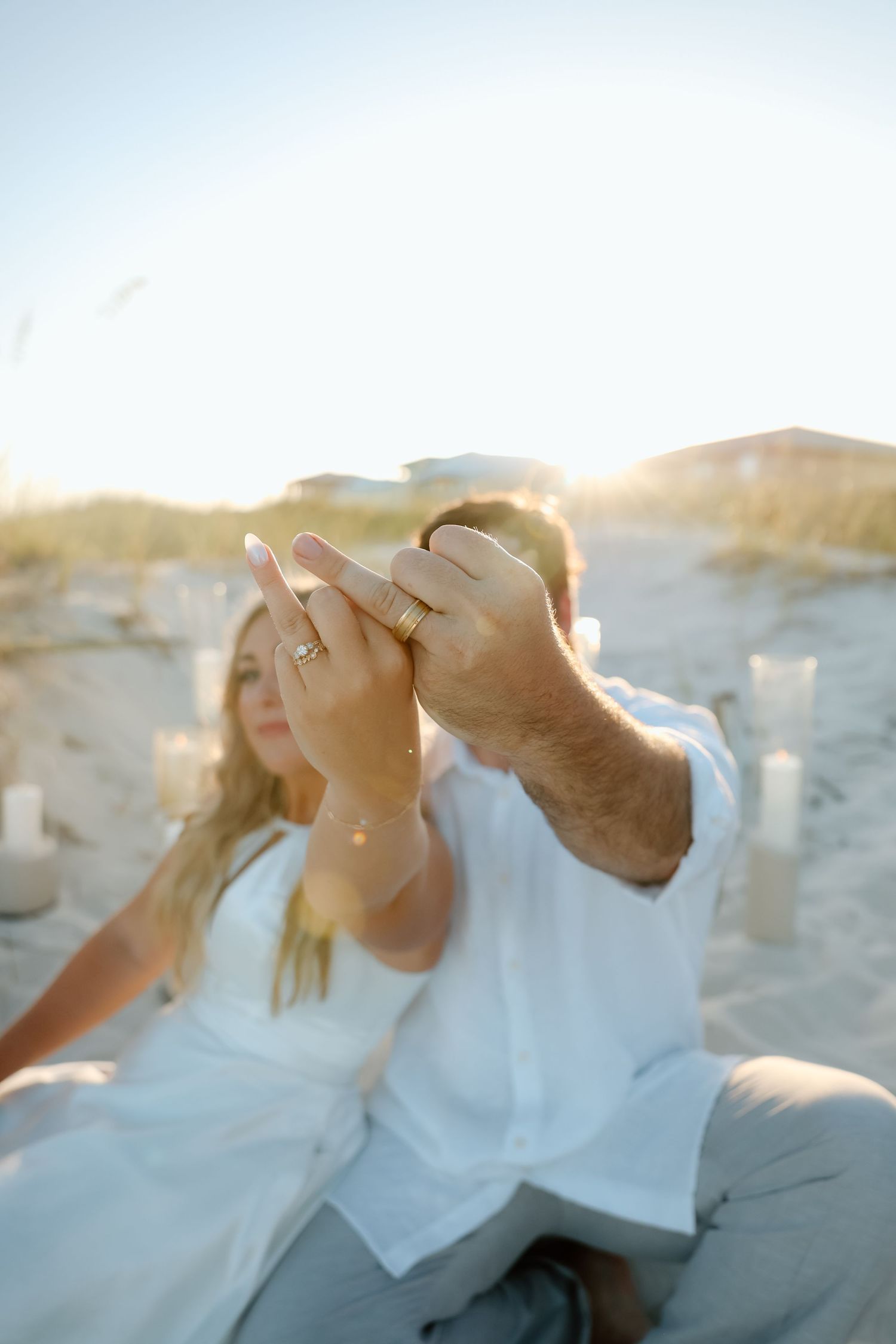 Newlywed couple holds up hands showing wedding rings together on sunlit beach at golden hour.