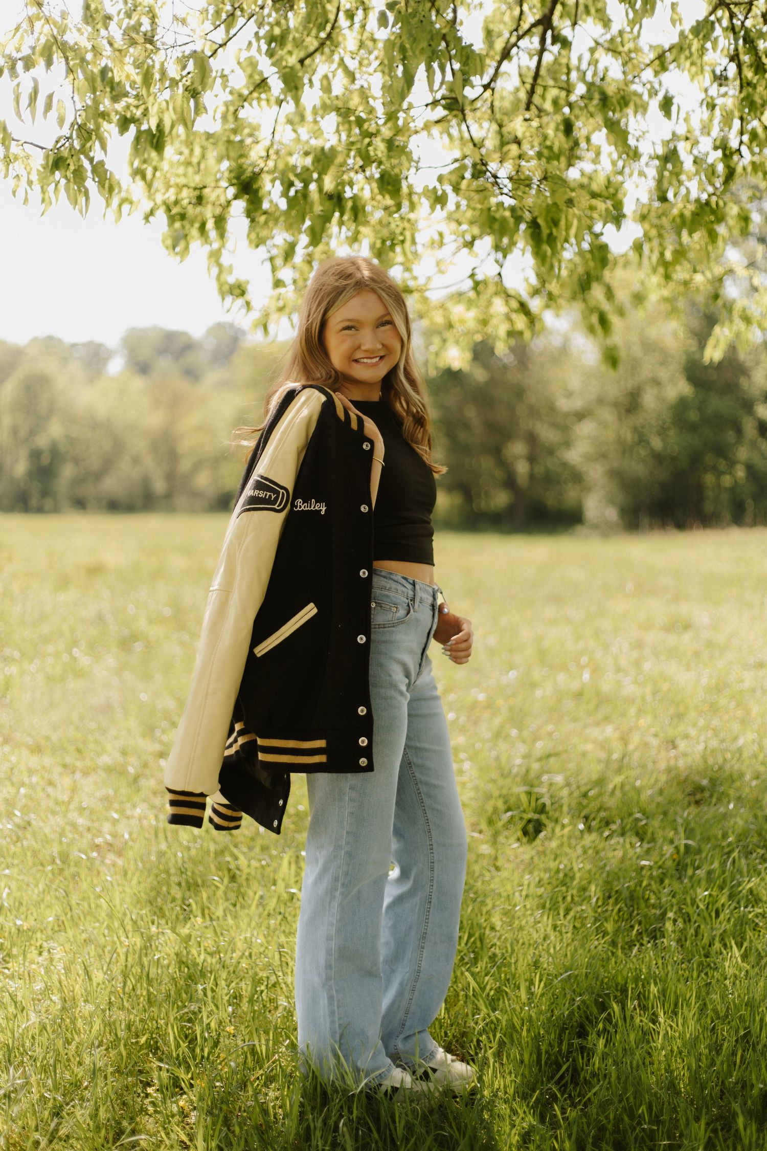 Smiling teenage girl in varsity jacket and wide-leg jeans poses in sunlit green meadow under tree.